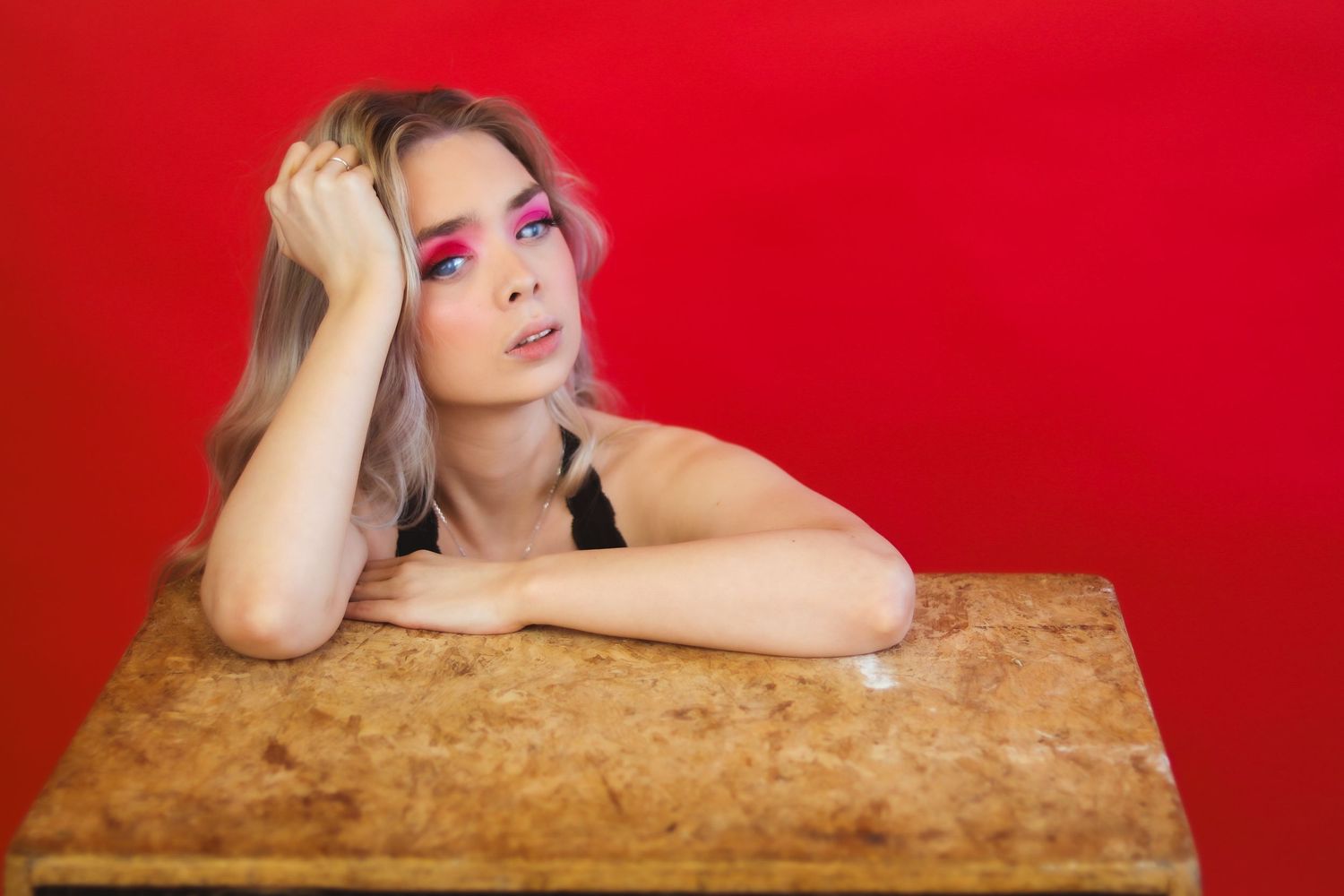 Young woman with bold pink eye makeup resting her head on her hand at a wooden table against red background.