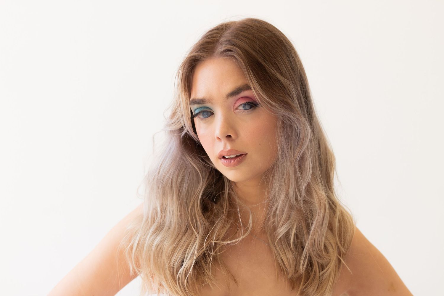 Young woman with wavy blonde hair and colorful eye makeup poses against a white background.