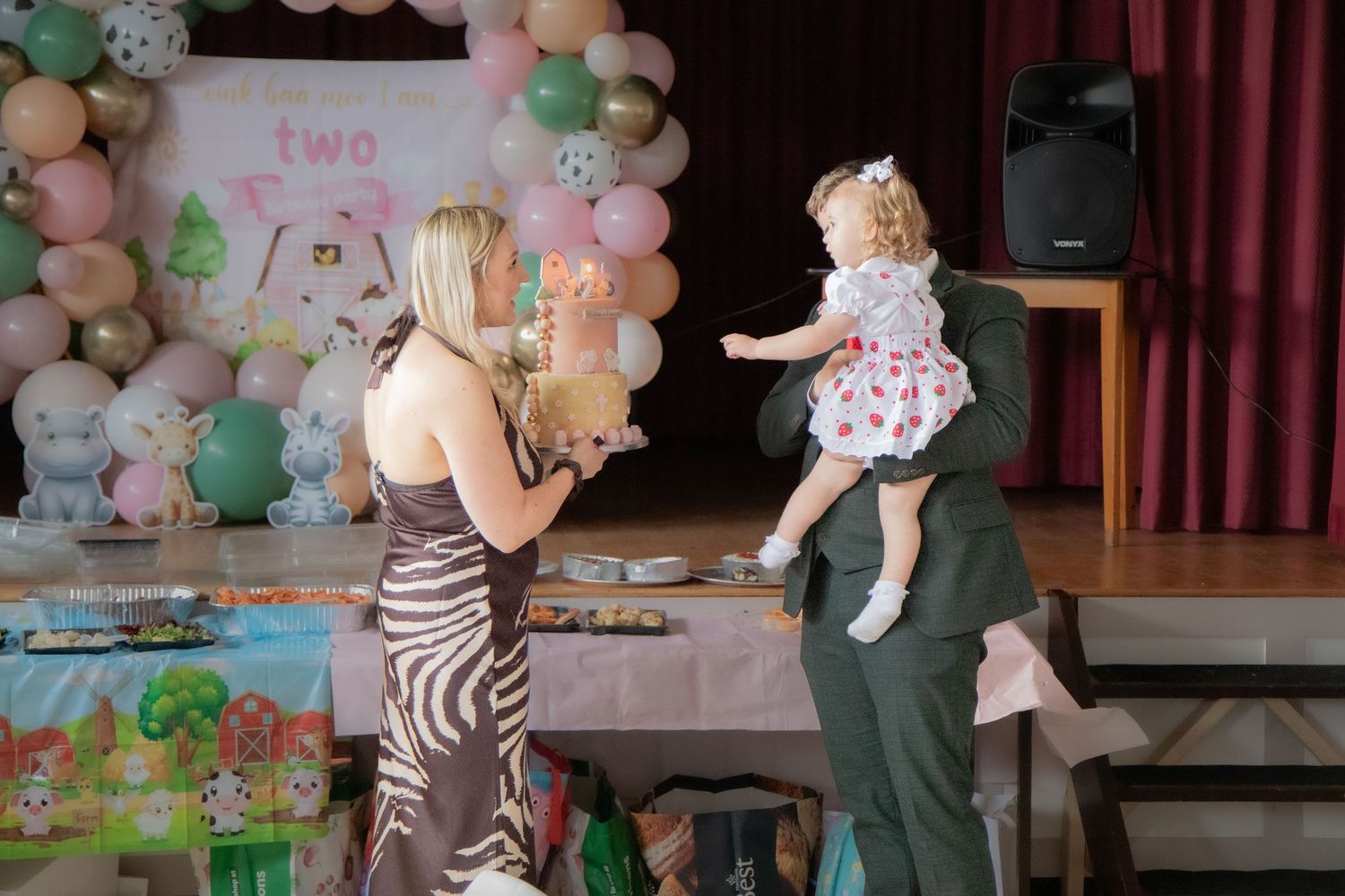 Woman presenting a birthday cake to a toddler girl in a floral dress at a colorful second birthday party.