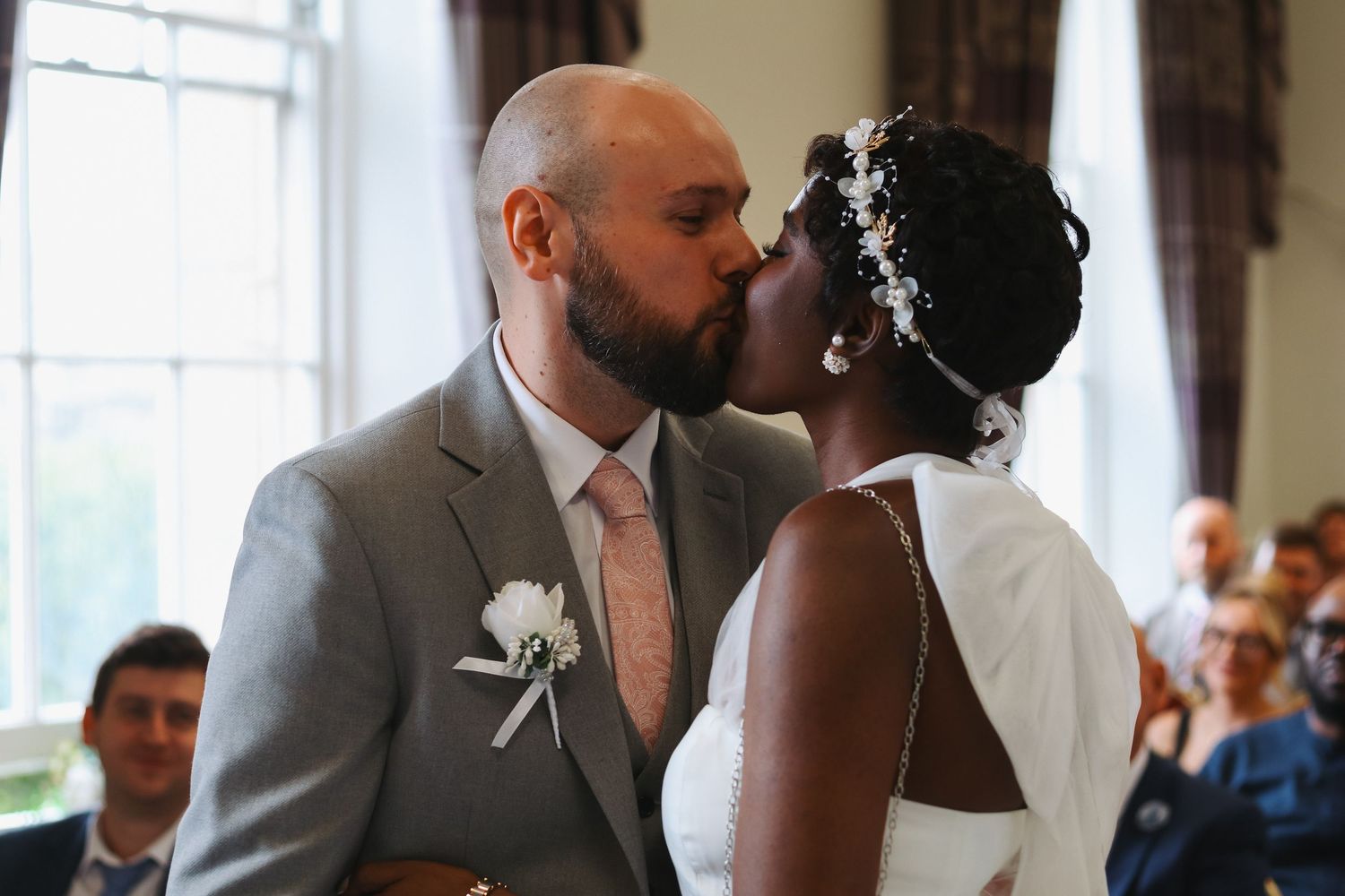 Interracial couple sharing first kiss at wedding ceremony, groom in gray suit, bride in white dress with floral headpiece.