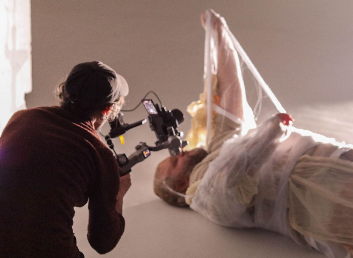 Photographer filming a wrapped sculptural figure lying on a white studio floor with professional camera equipment.
