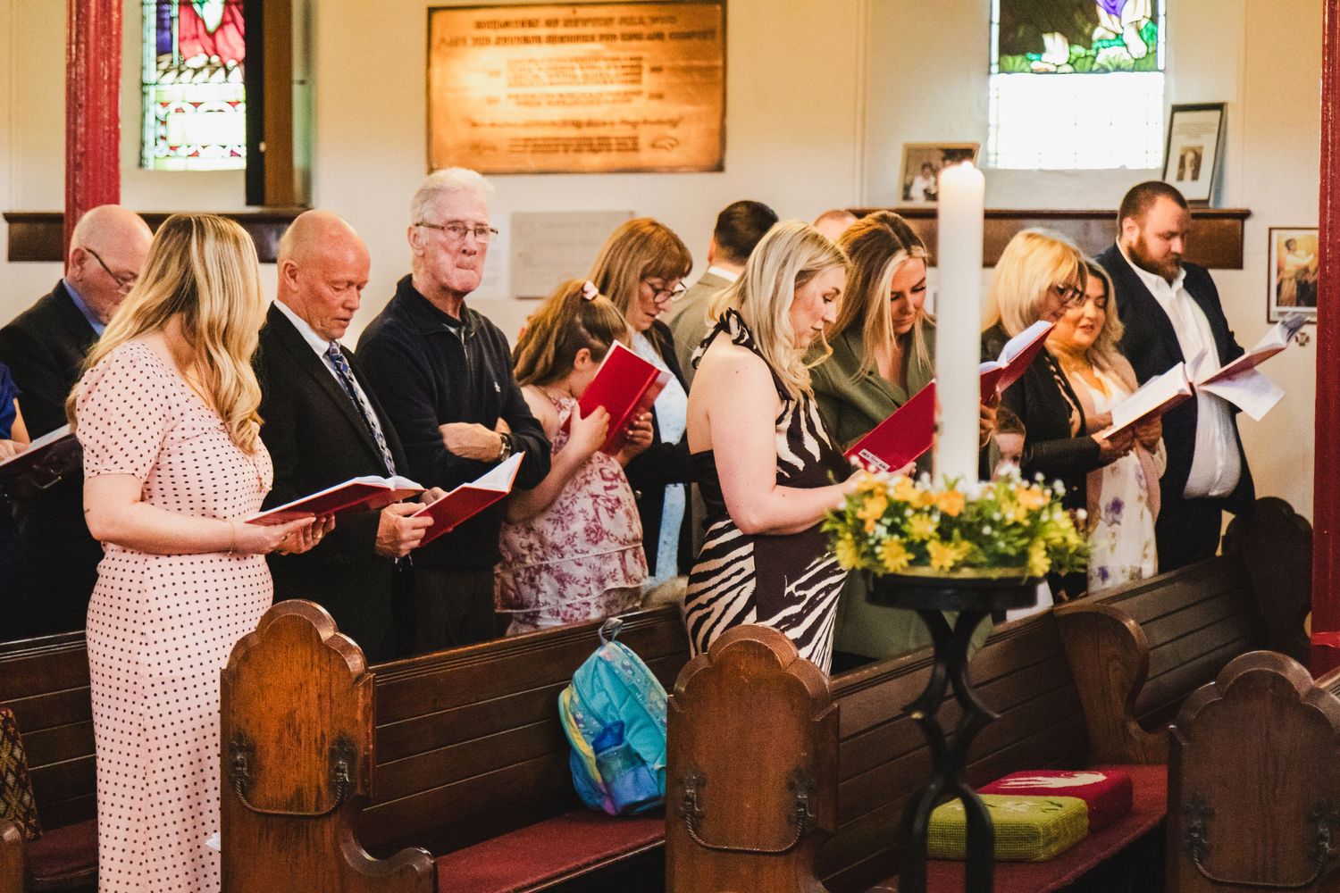 Congregation standing in wooden church pews reading hymn books during a religious ceremony service.