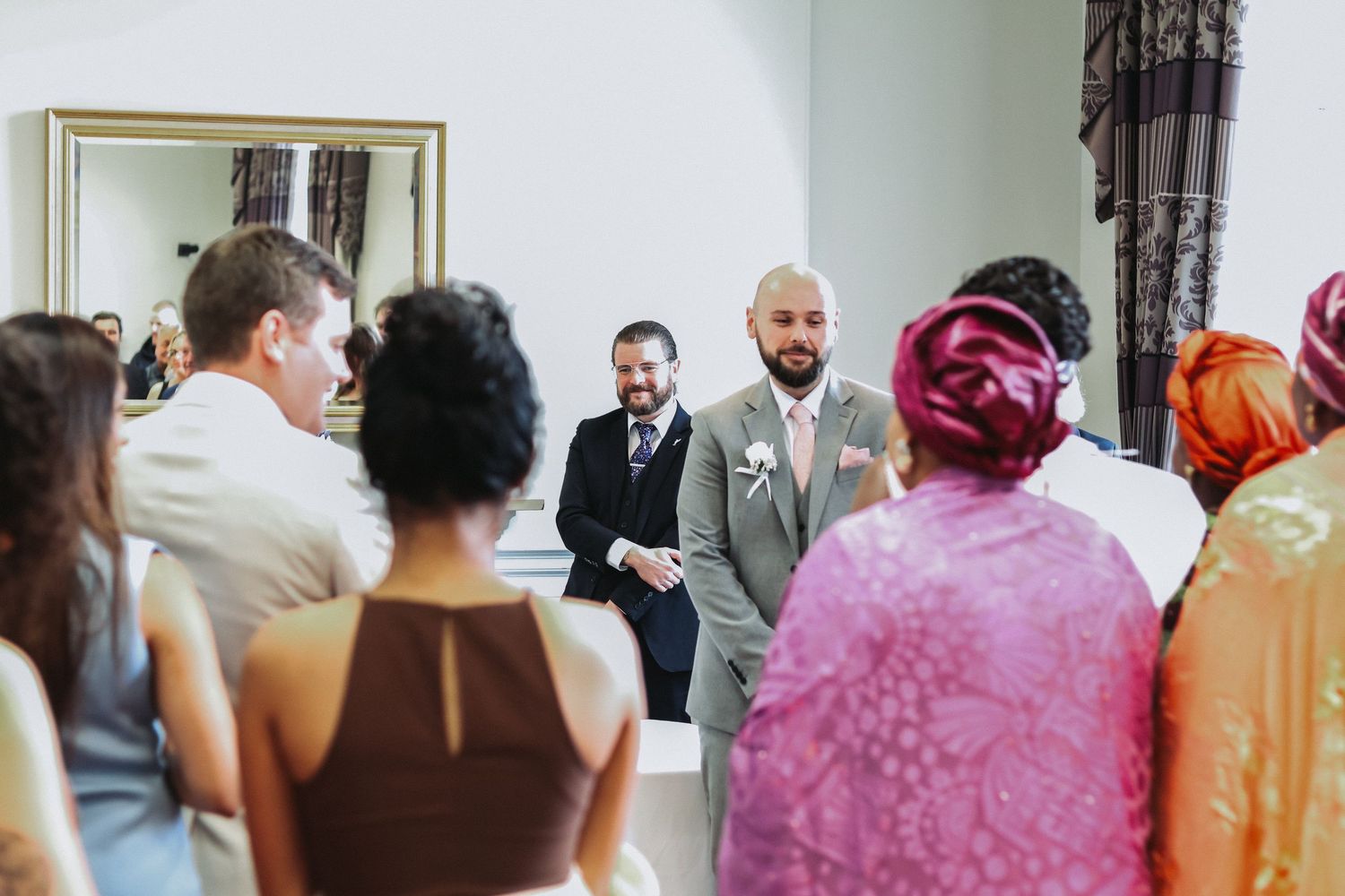 Groom stands smiling at the altar as wedding guests are seated in a bright elegant room.