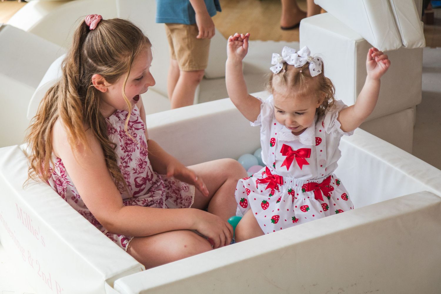 Young girl smiling at excited toddler in white play area wearing red bow dress.