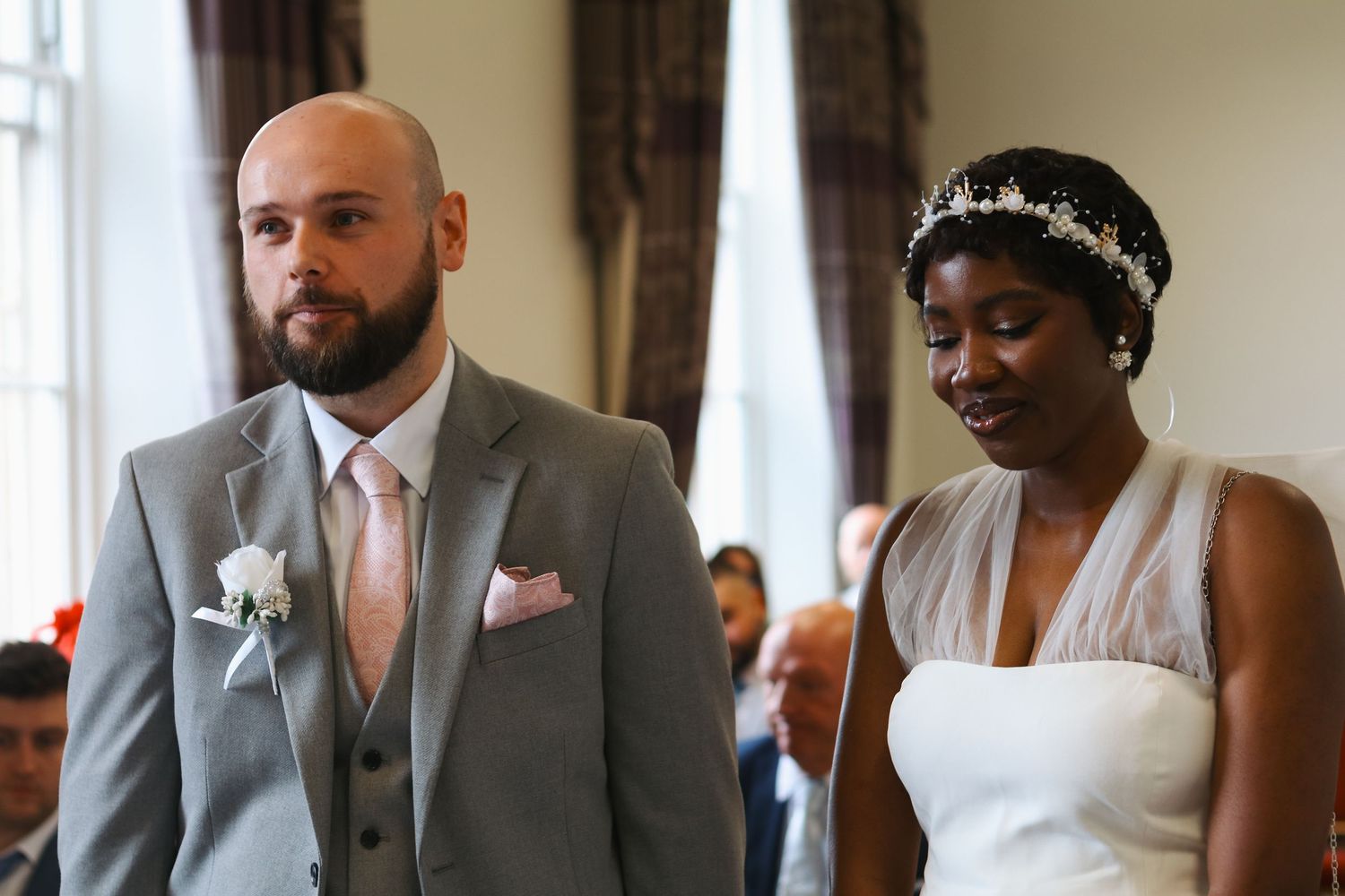Bride and groom standing together during wedding ceremony, groom in gray suit, bride in white gown with floral headpiece.
