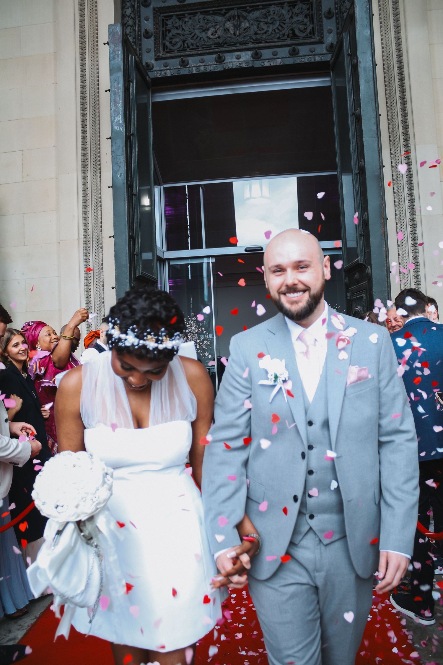 Newlywed couple smiling as colorful heart confetti falls outside wedding venue, bride in white dress, groom in gray suit.