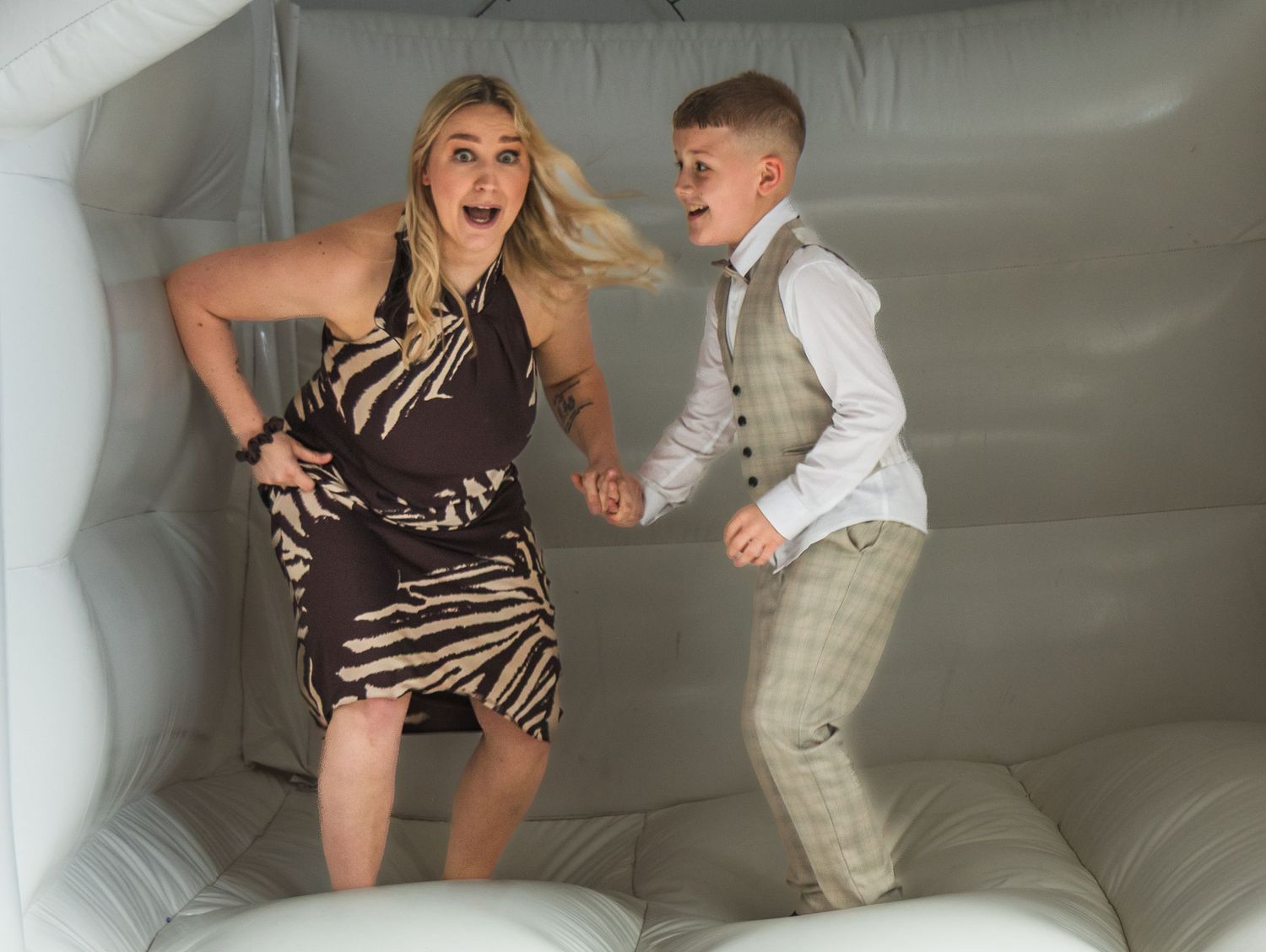 Woman and boy jumping joyfully in a white inflatable bounce house, dressed in elegant party attire.