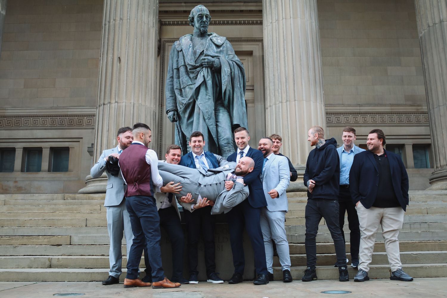 Group of men in suits lifting one person outside a neoclassical building with large columns.