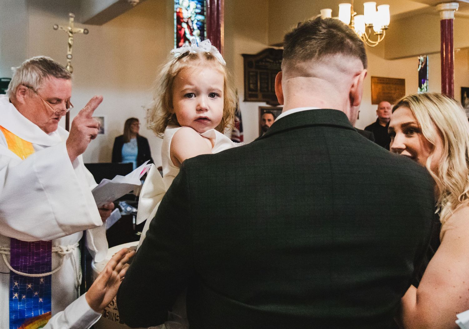A young flower girl being held at a church wedding ceremony while a priest performs the service.