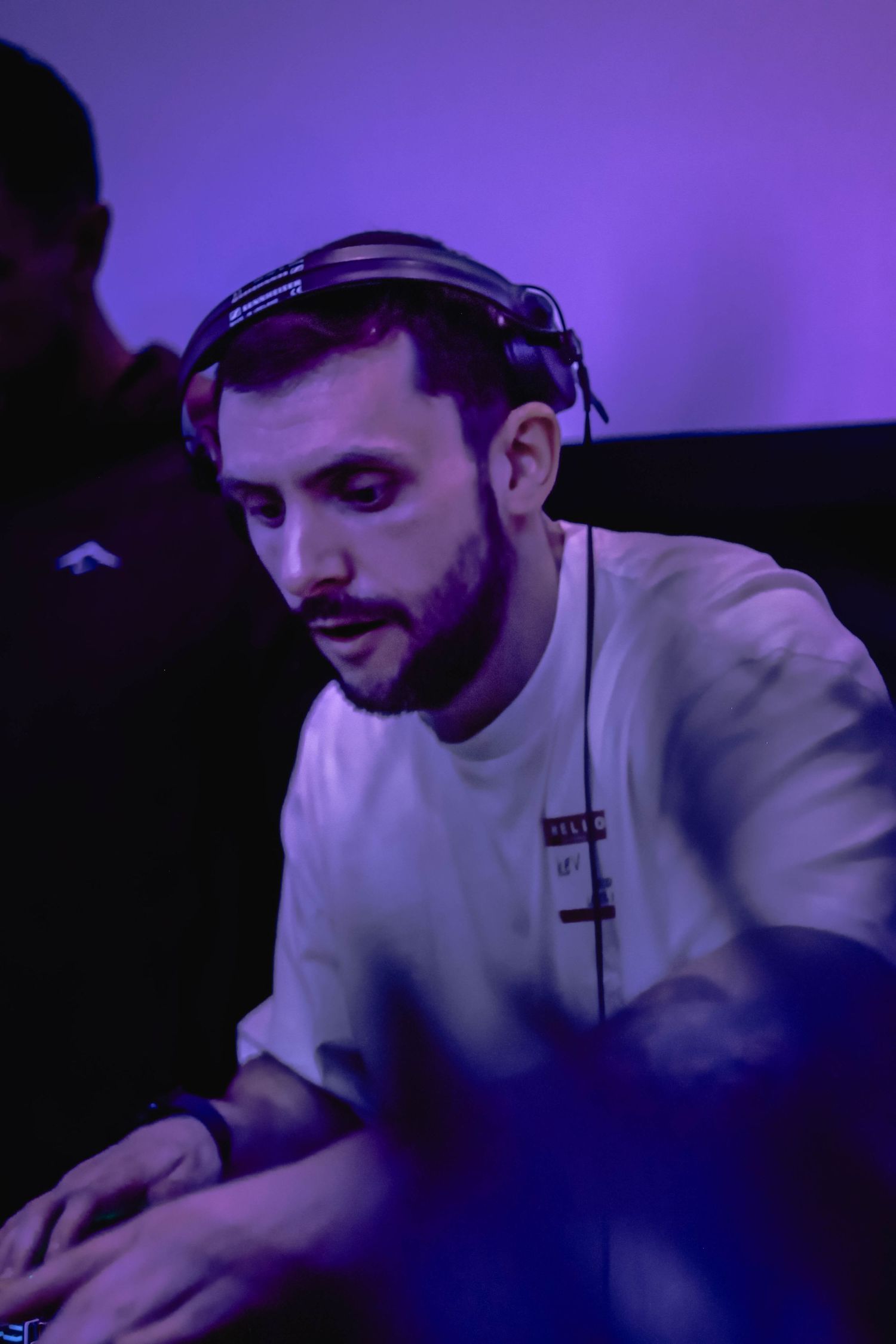 A male DJ with headphones and beard wearing a white shirt performs under purple lighting in a dark club setting.