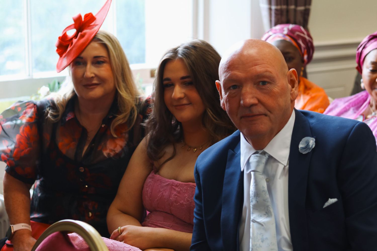 Two women and a man in formal wedding guest attire smile together at a wedding reception indoors.