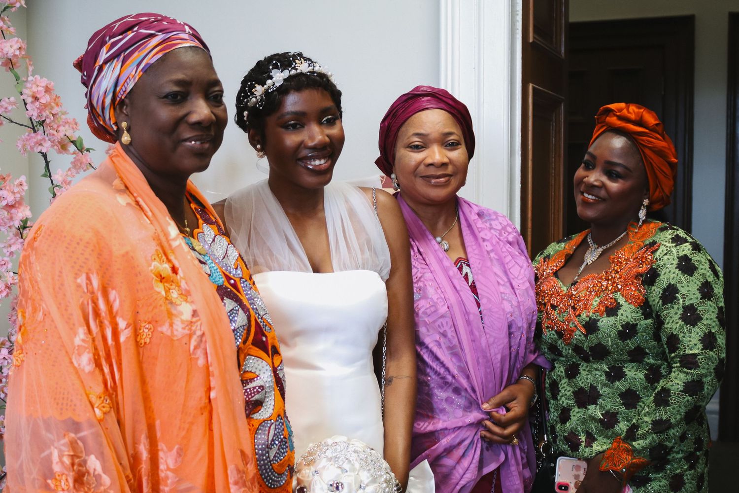 A bride in a white gown poses with three women in vibrant African attire at a wedding celebration.