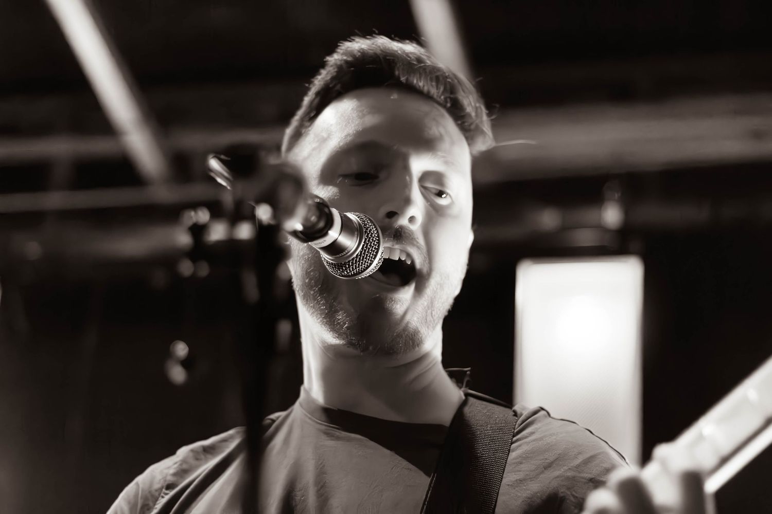 Young male vocalist singing passionately into microphone while playing guitar in black and white photo.