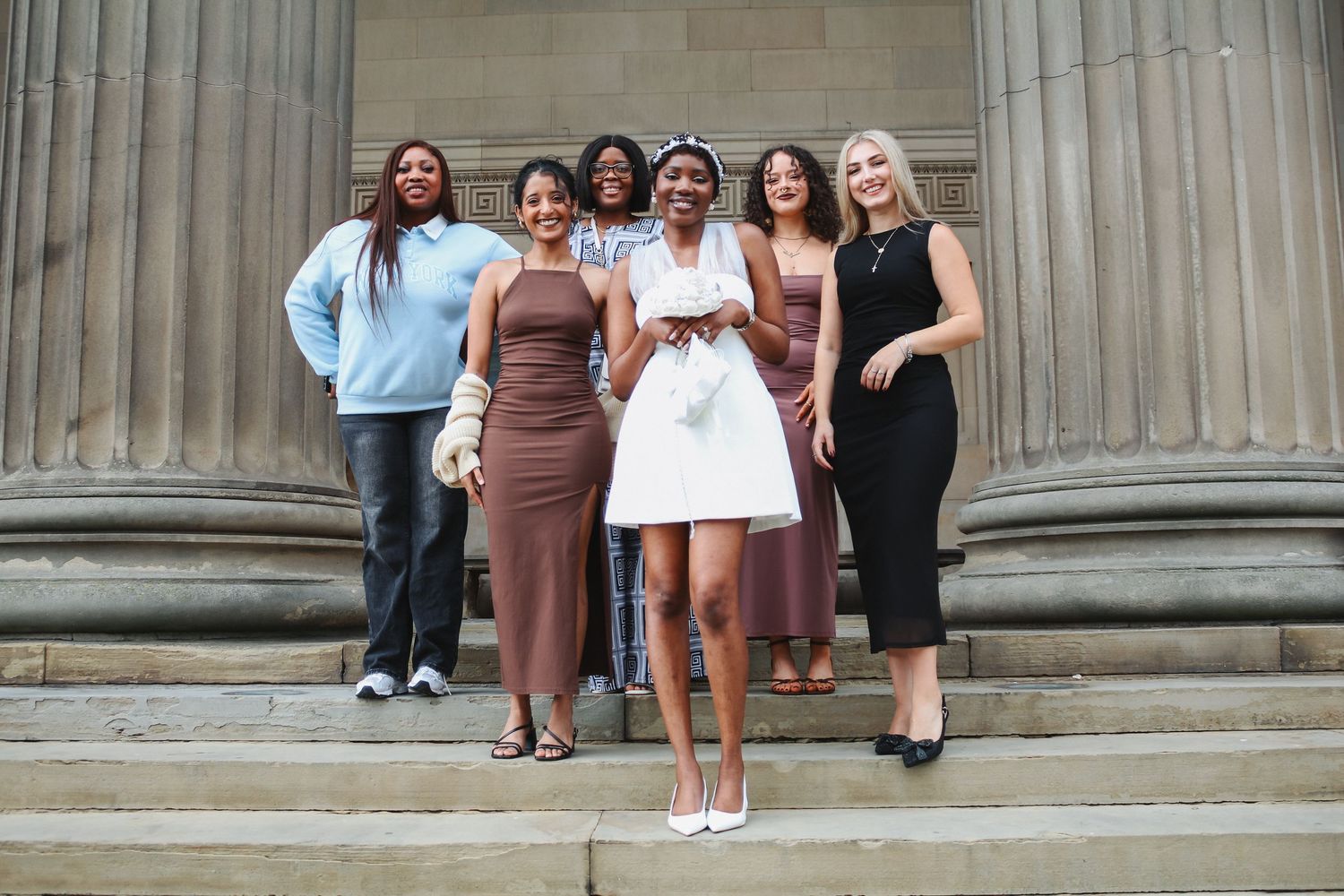 Bride in white mini dress poses with five bridesmaids in brown dresses on stone steps.