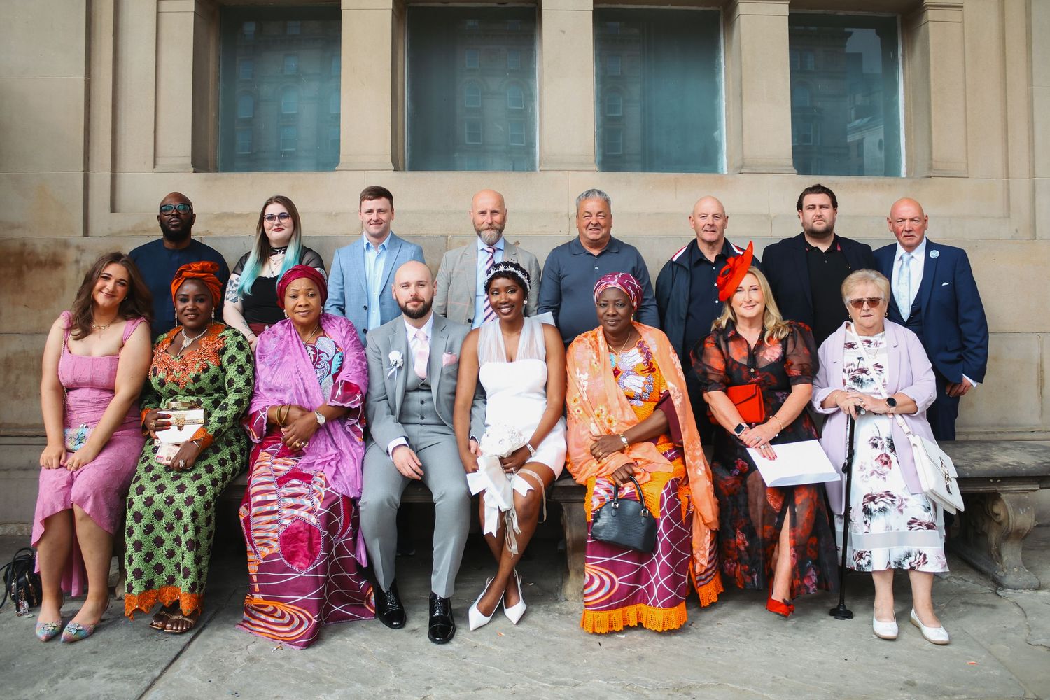 Diverse group of people in colorful traditional and formal attire posing together on steps of a historic building.