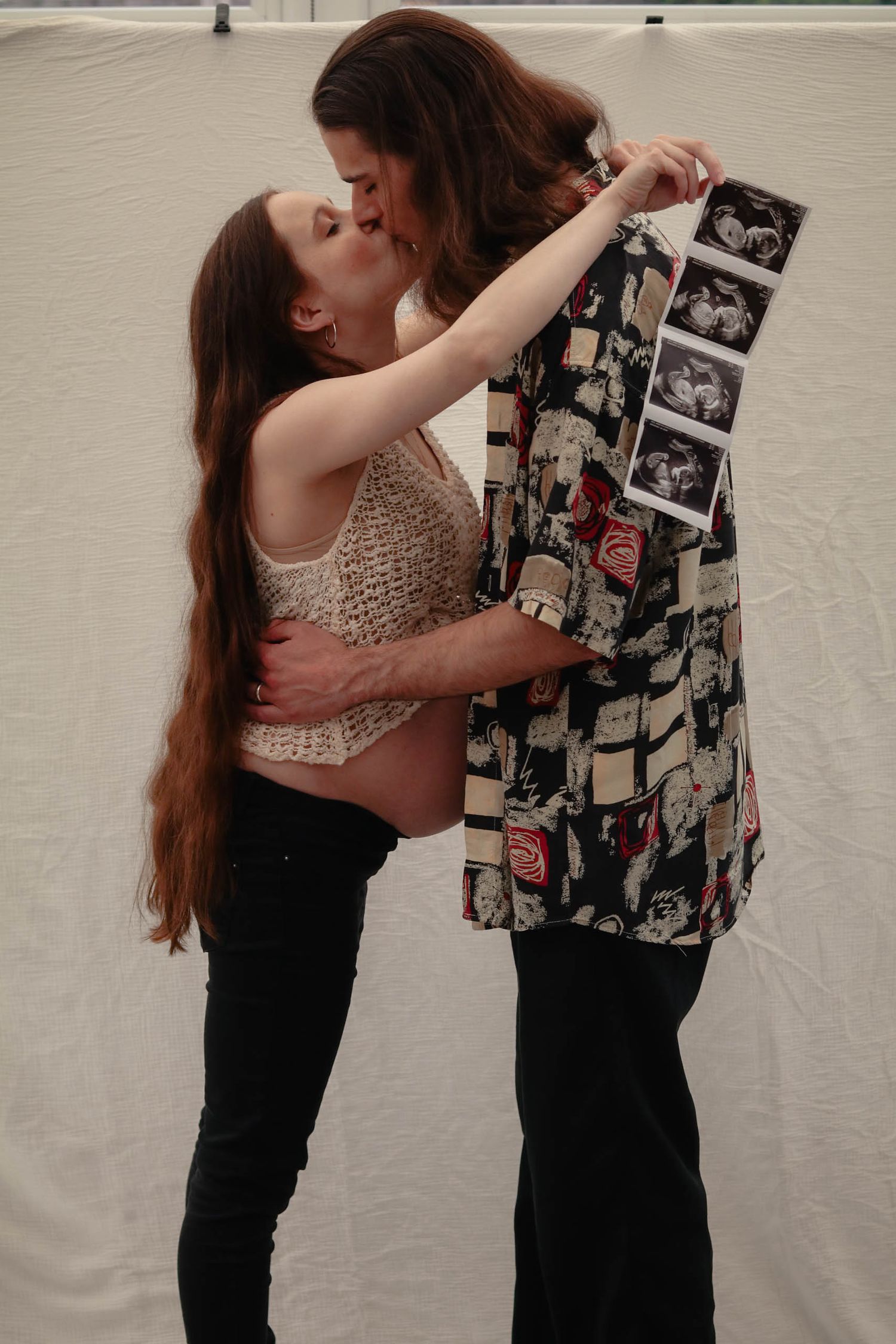 Two women sharing a kiss while holding ultrasound photos, celebrating a pregnancy announcement together.