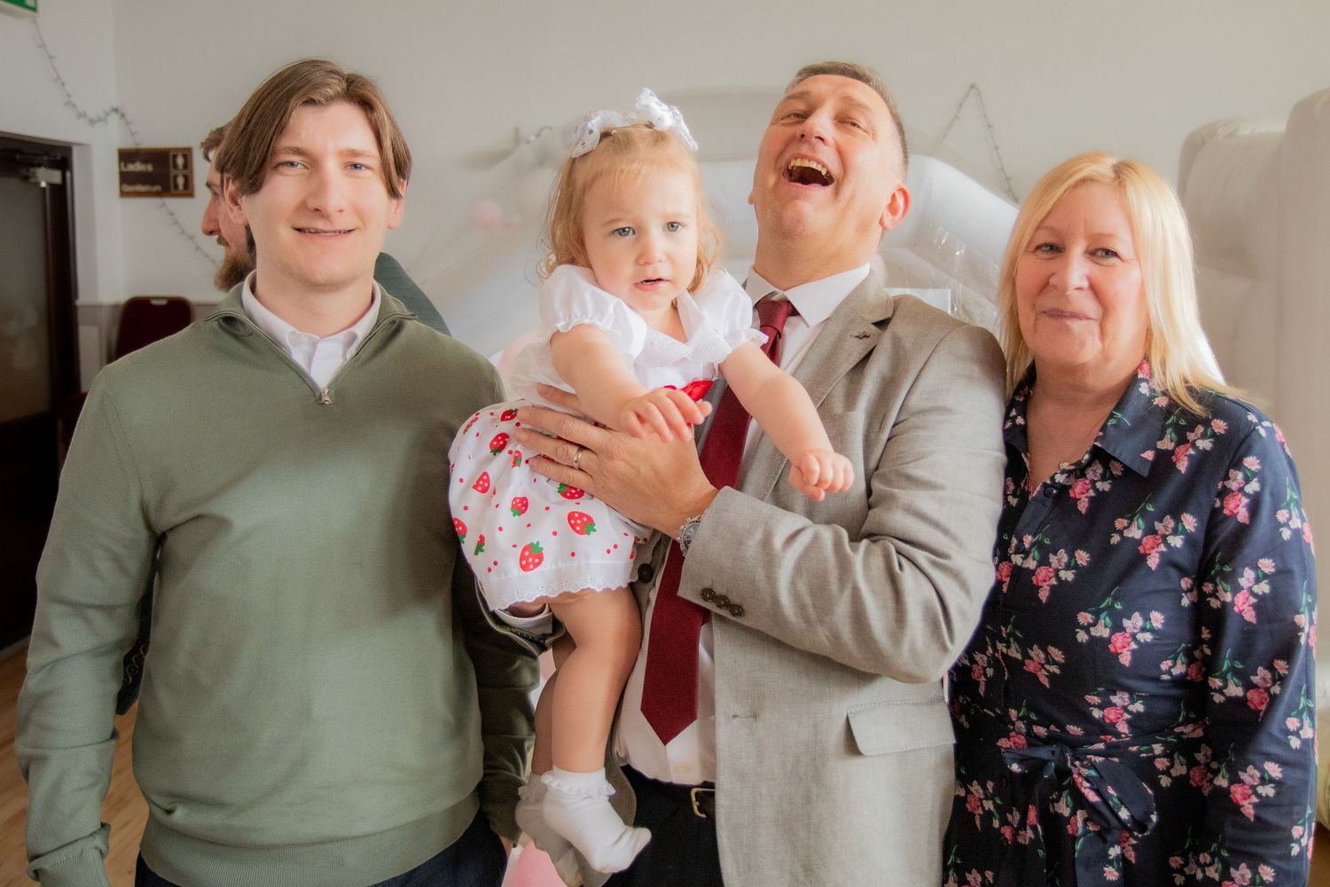Family group smiling and laughing together at a celebration, with a man holding a baby girl in a white dress.