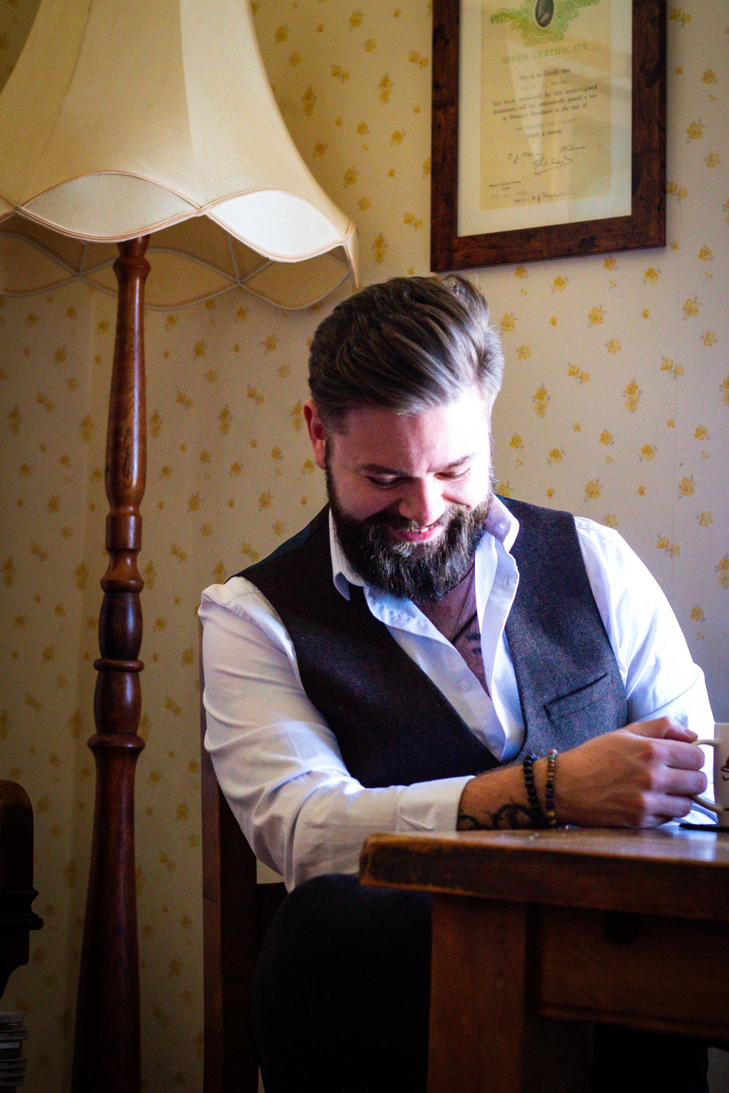 Bearded man in a grey vest and white shirt sitting at a wooden desk in a vintage-styled room.
