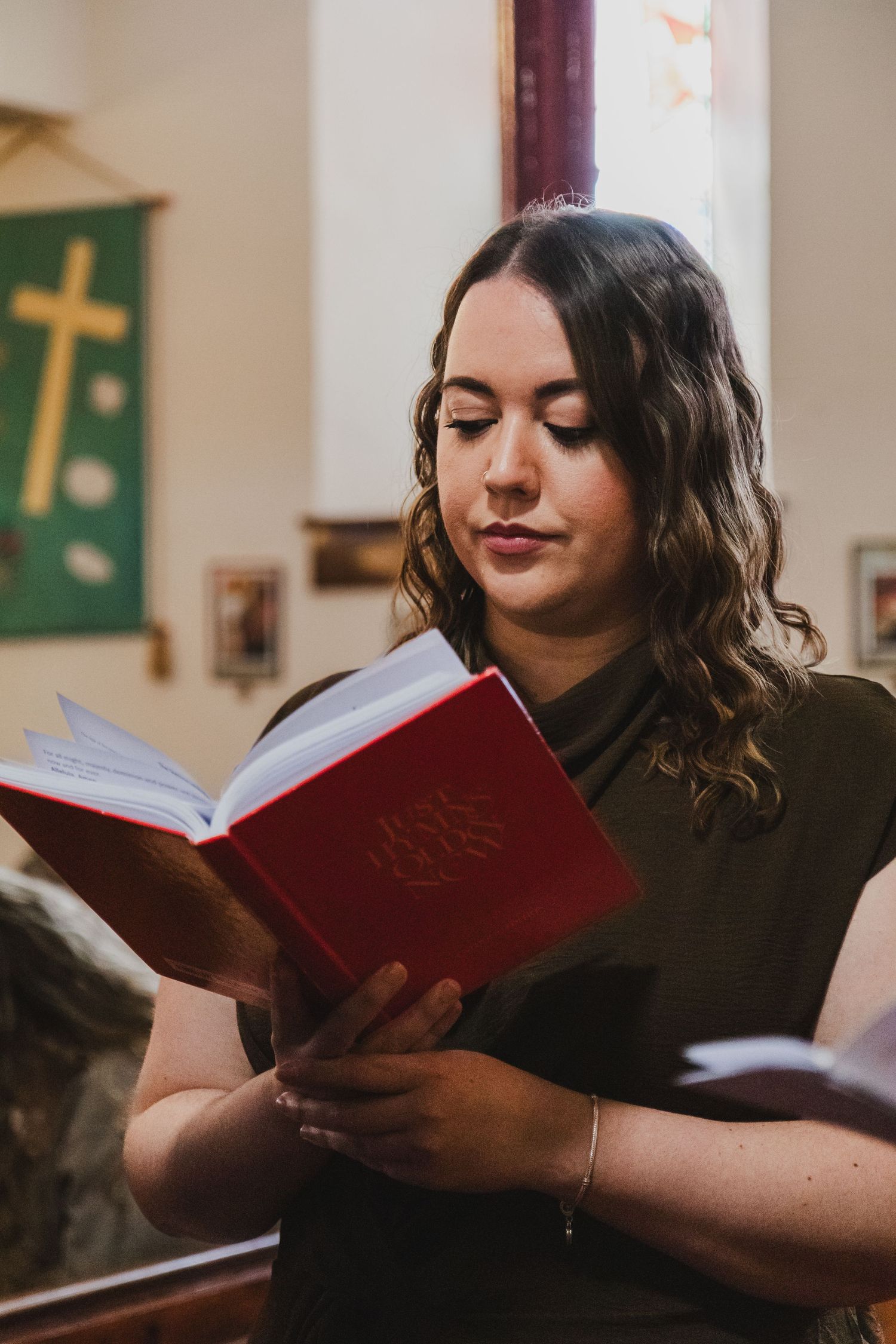 Young woman reading a red hymnal book in a church with a golden cross banner visible in the background.