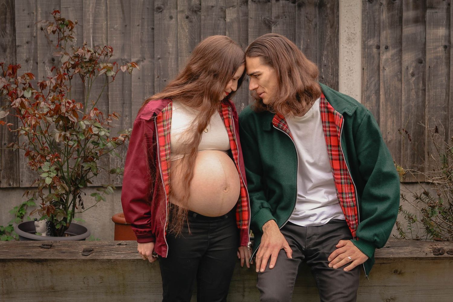 Pregnant couple touching foreheads outdoors, wearing matching plaid jackets, sharing an intimate moment by a wooden fence.