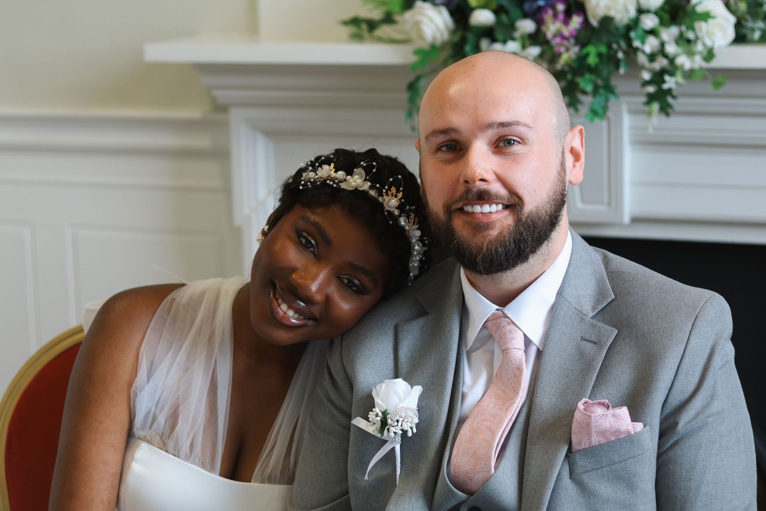 Interracial newlywed couple smiling together, bride wearing floral tiara and groom in grey suit with boutonniere.