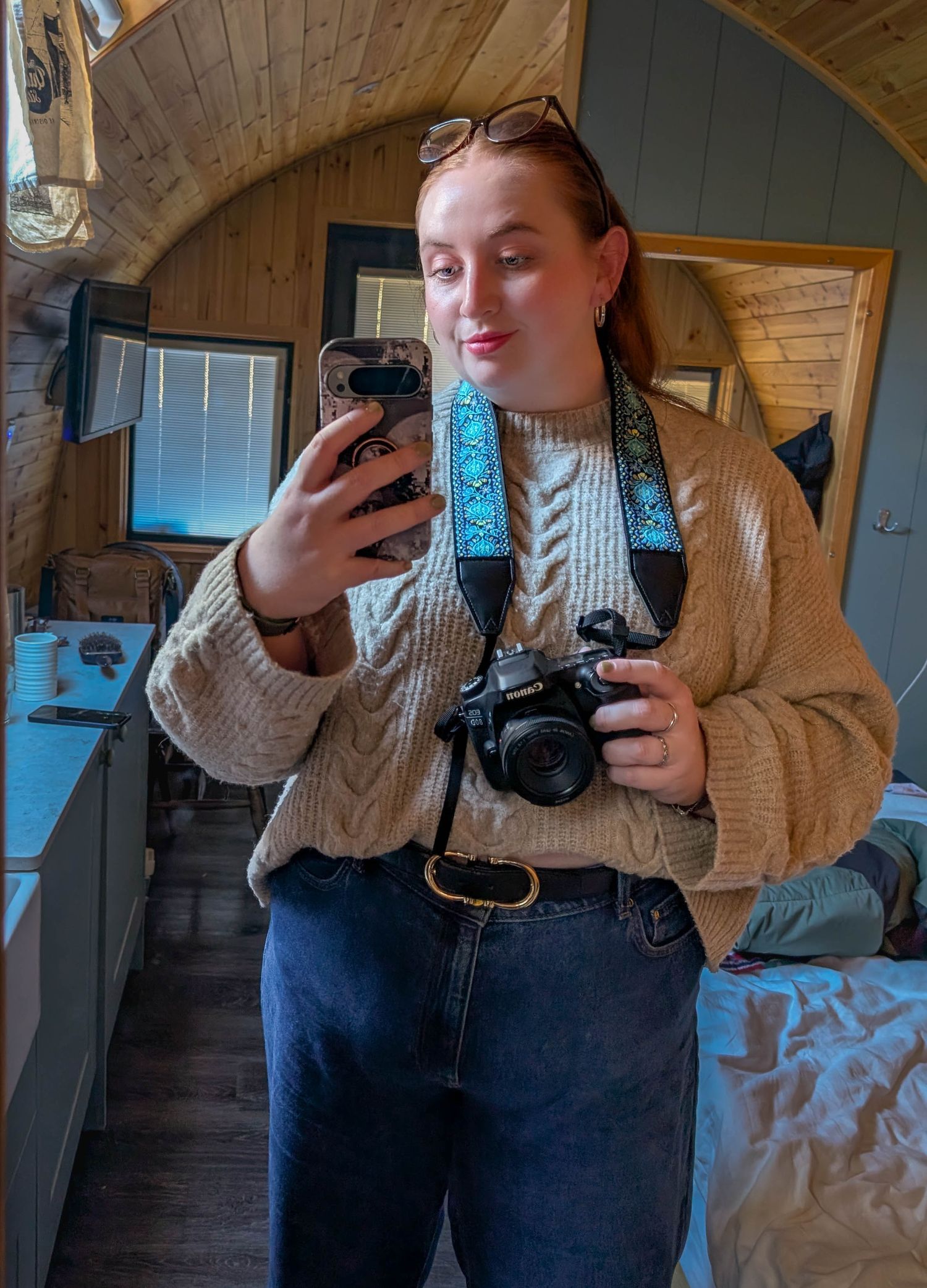 Young woman in cable-knit sweater and jeans takes mirror selfie holding DSLR camera with blue strap in rustic wooden interior.