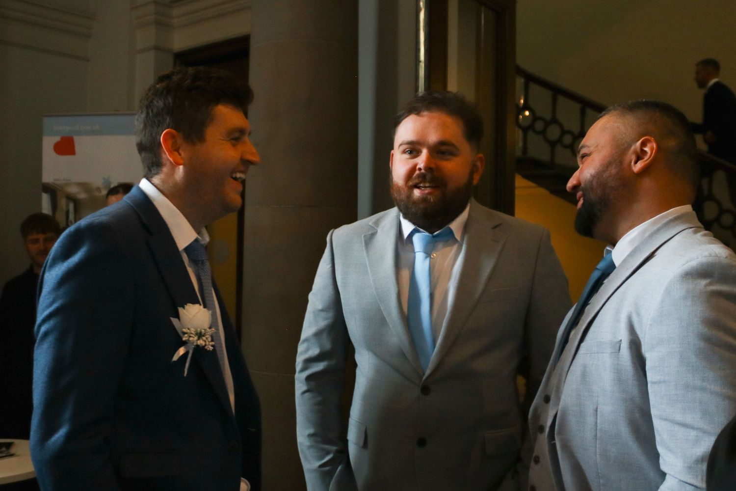 Three men in suits engaged in conversation at a formal event or wedding reception indoors.