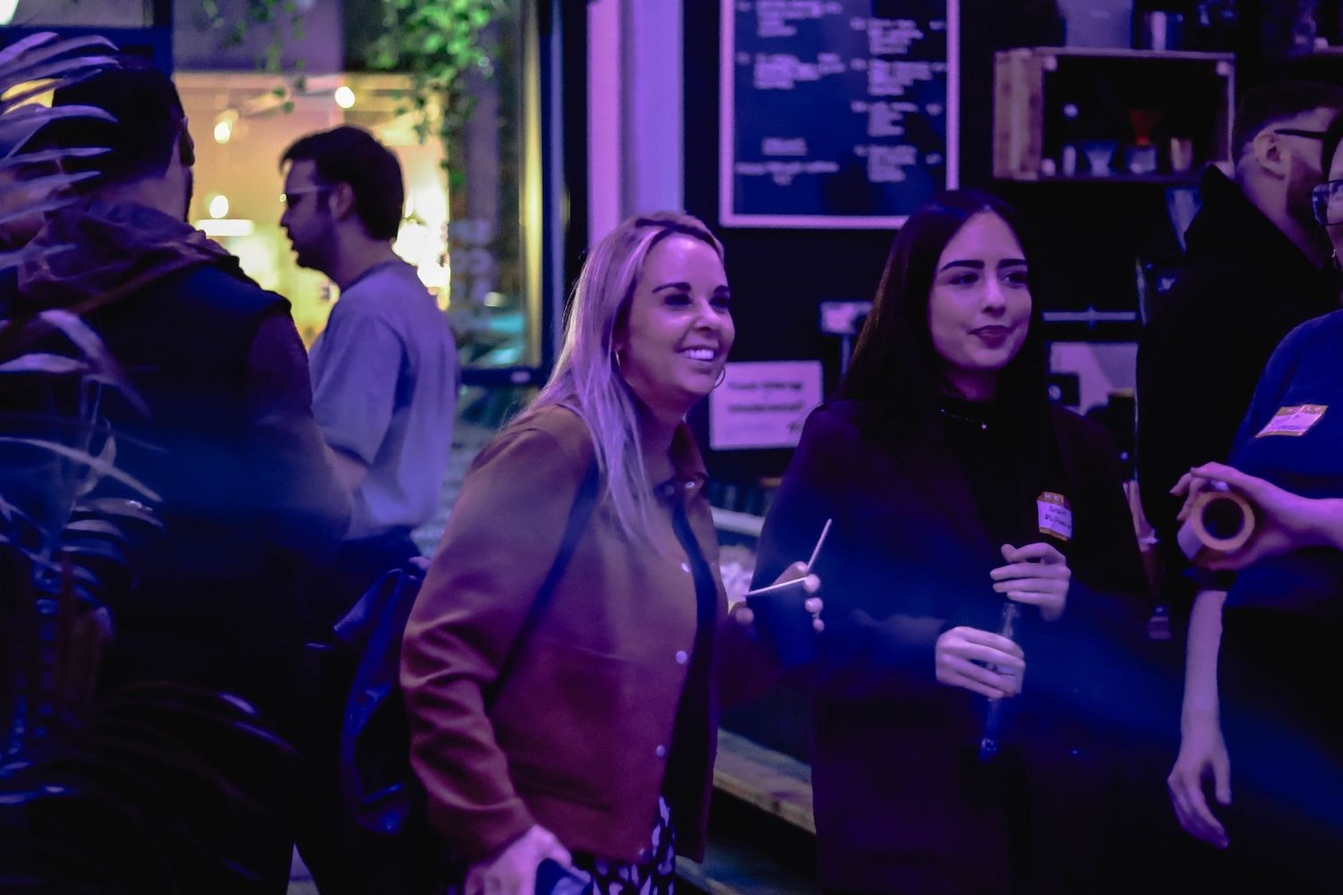 Smiling blonde woman in brown jacket socializing at a dimly lit bar event with purple ambient lighting.