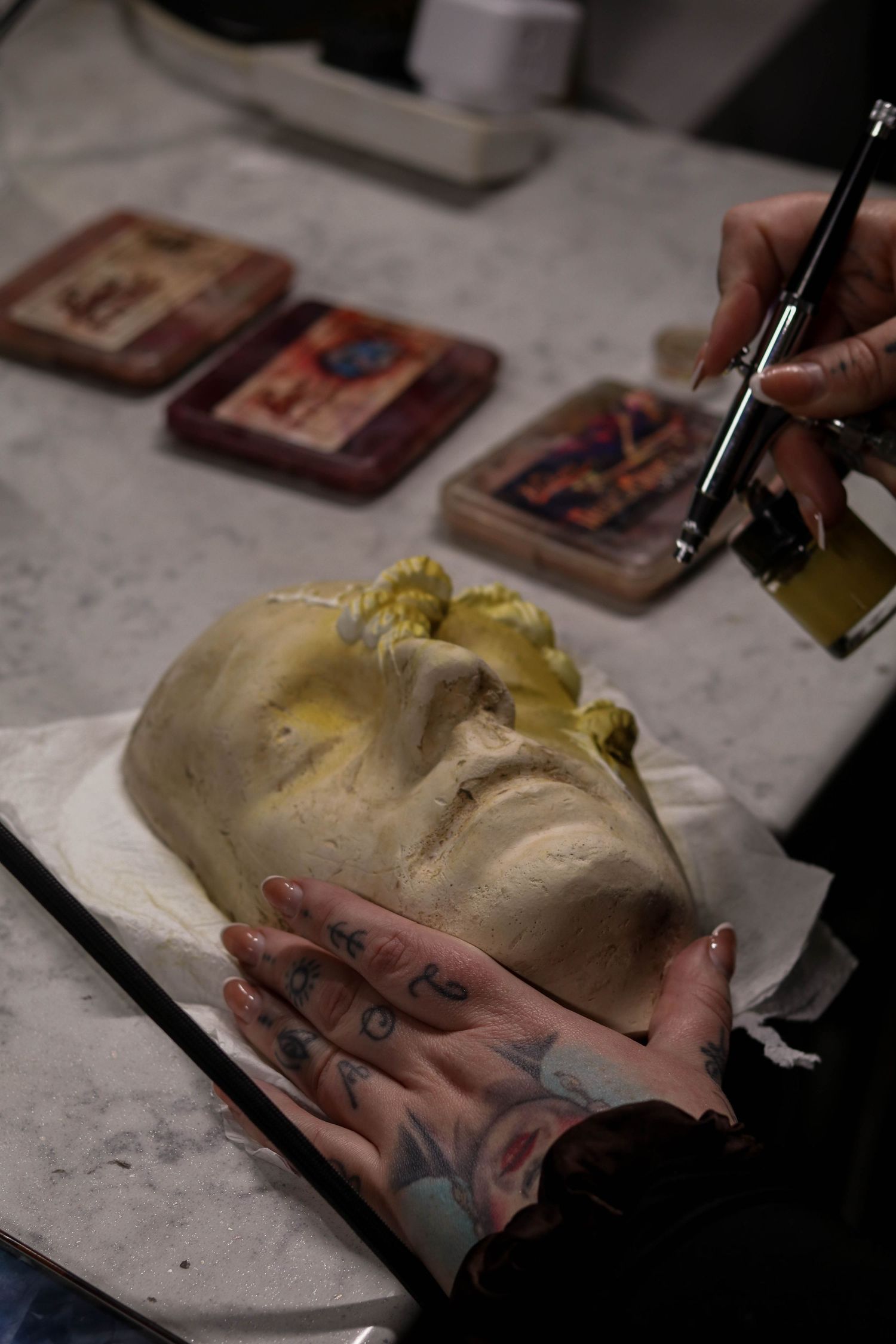 Tattooed hands hold a face cast sculpture while an airbrush applies yellow paint in a special effects makeup studio.