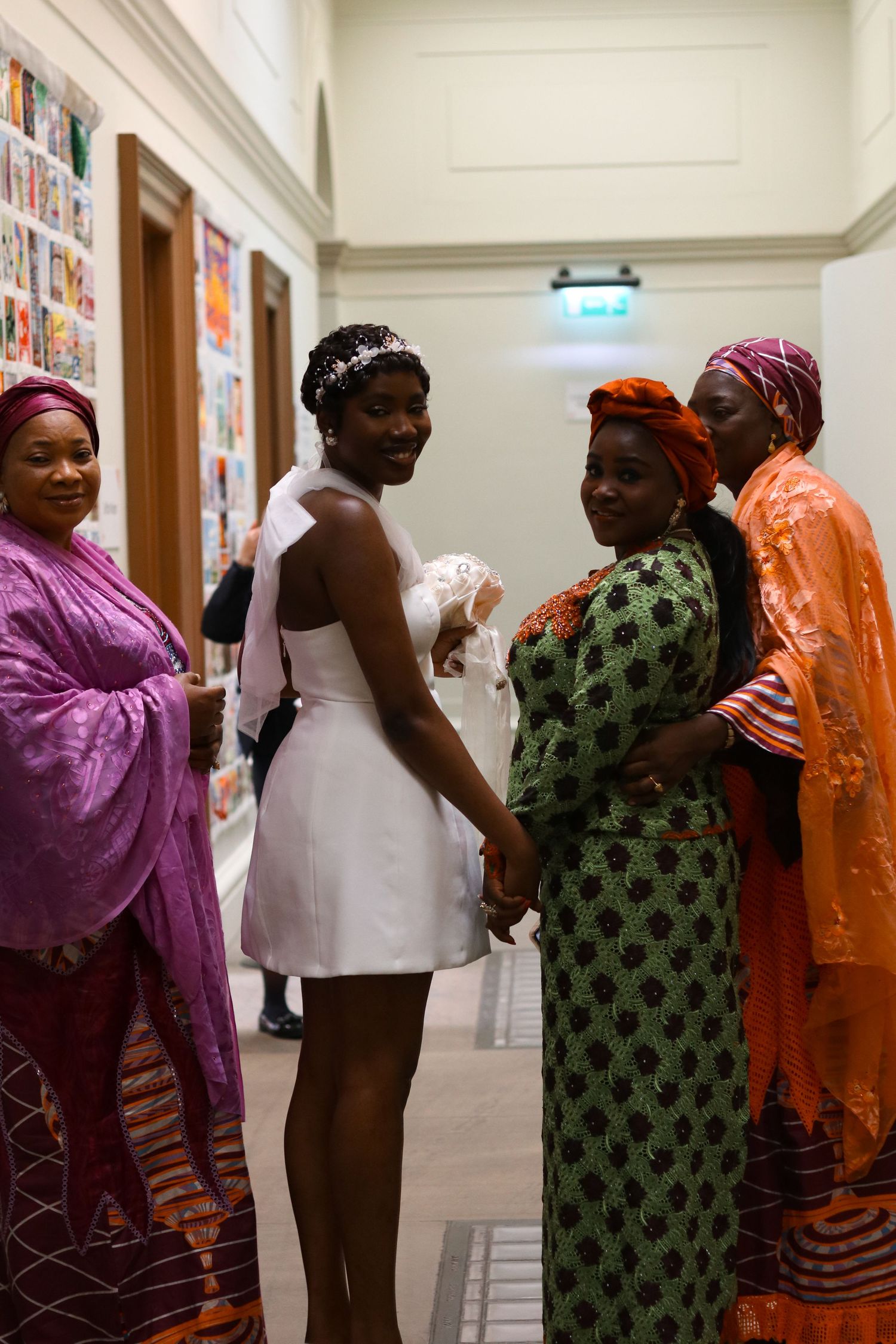 African women in colorful traditional attire pose with a bride in white dress and veil in an elegant hallway.