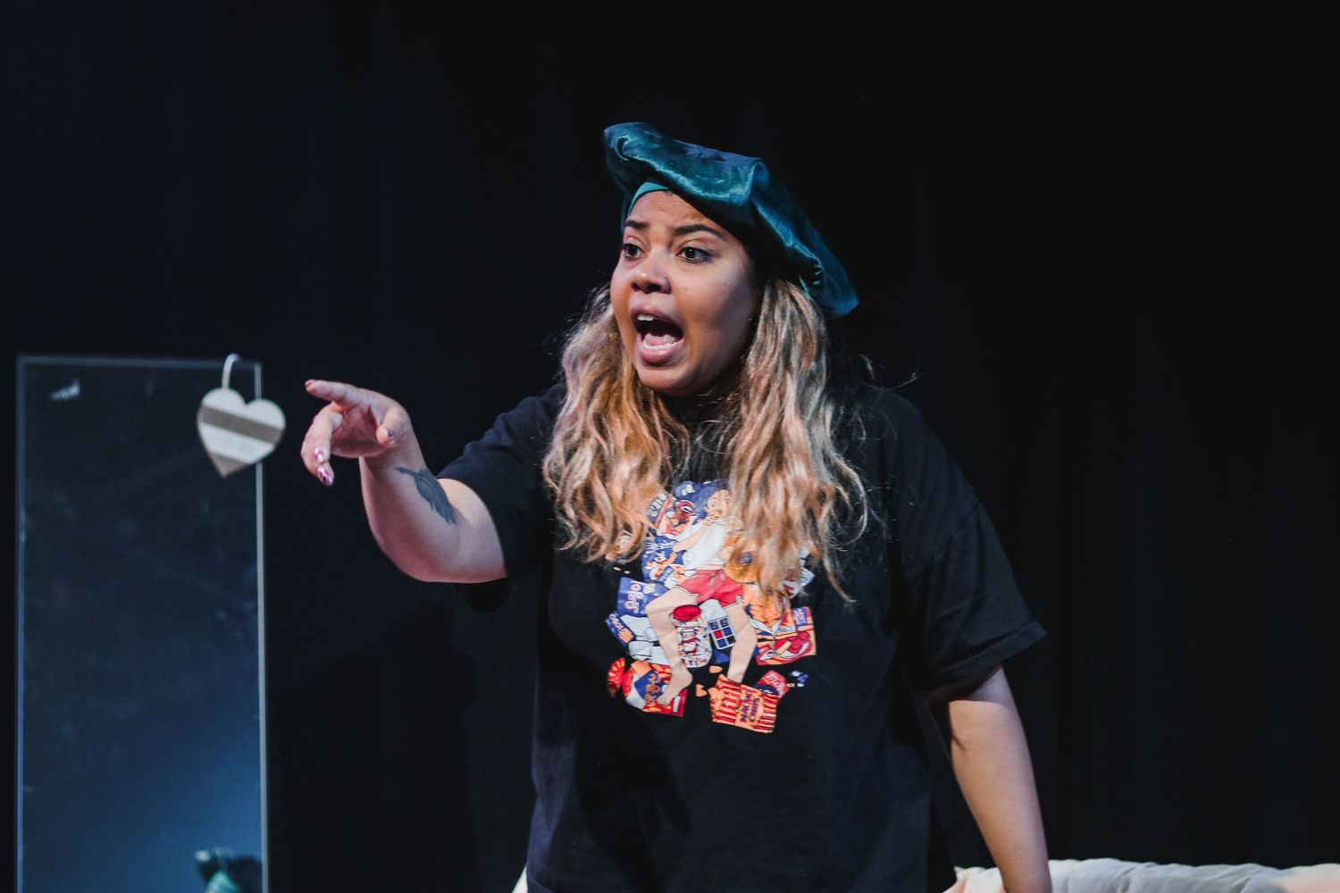 An expressive performer in a black graphic tee and teal cap points dramatically on a dark stage.