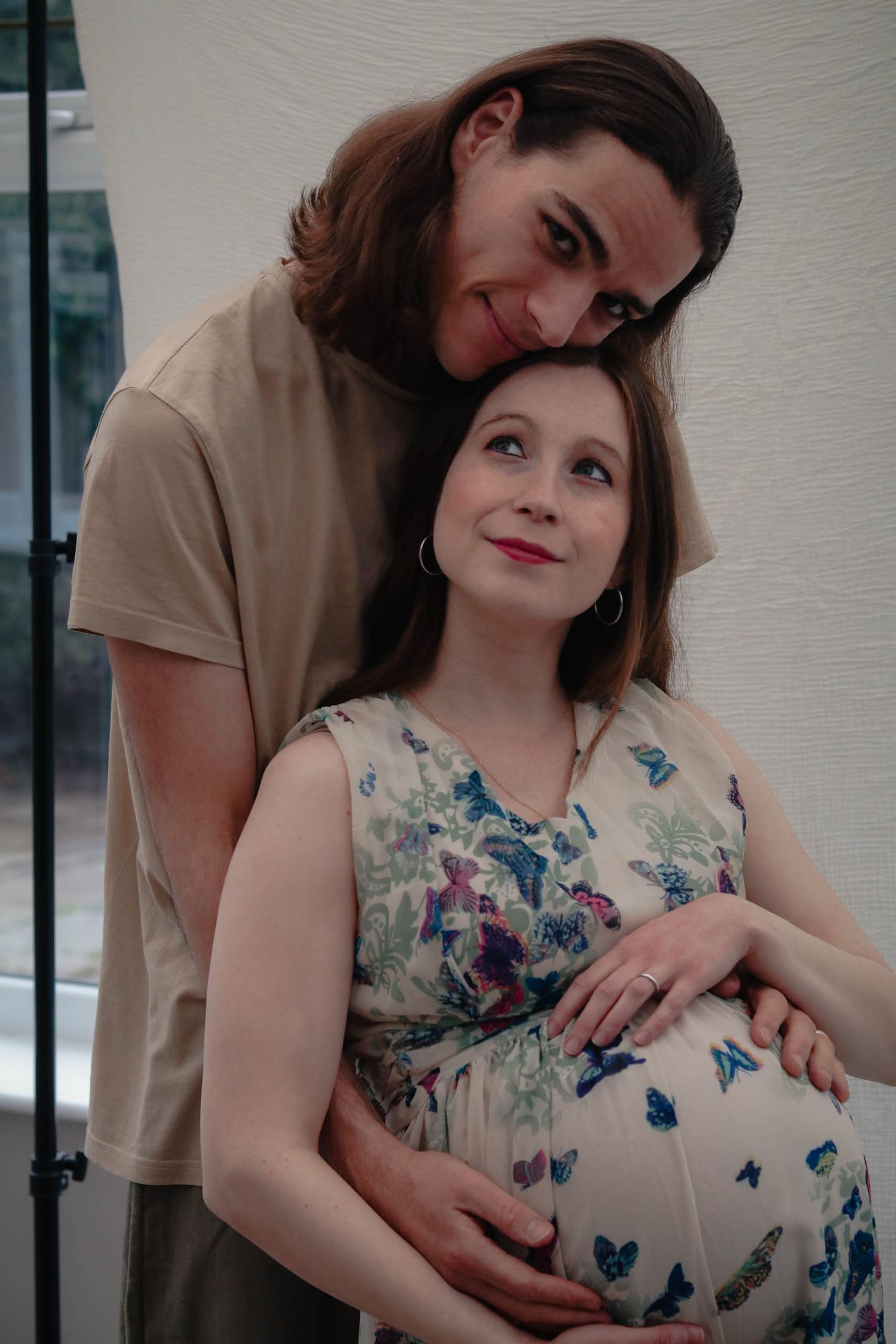 A pregnant woman in a floral dress cradles her baby bump while her partner embraces her from behind, smiling warmly.
