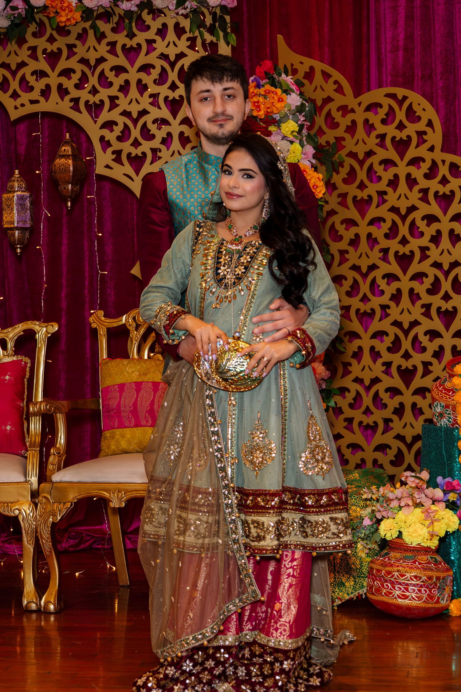Couple in traditional South Asian wedding attire posing at a decorated mehndi ceremony stage with floral backdrop.