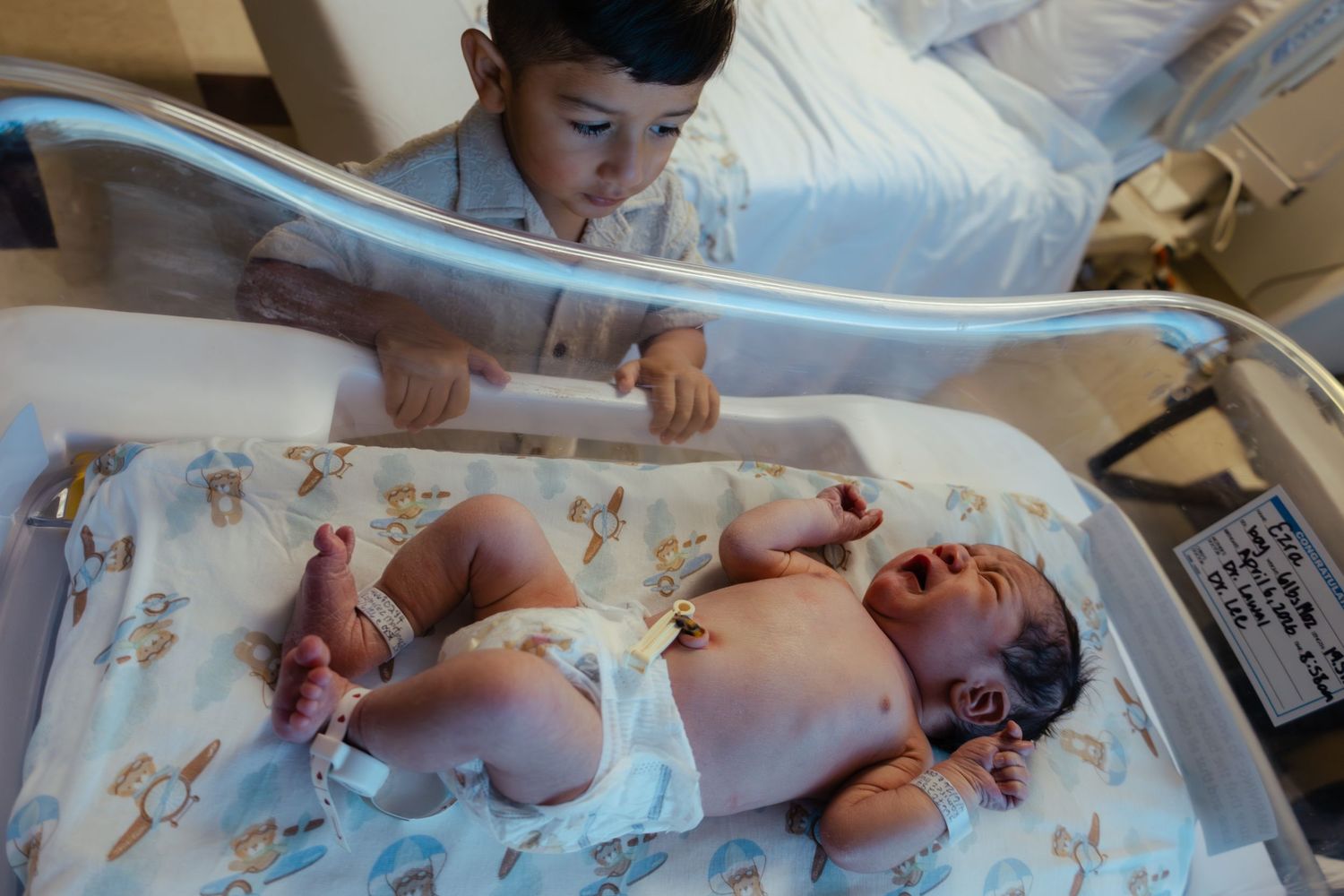Young boy gazes curiously at his newborn sibling lying in a hospital bassinet.