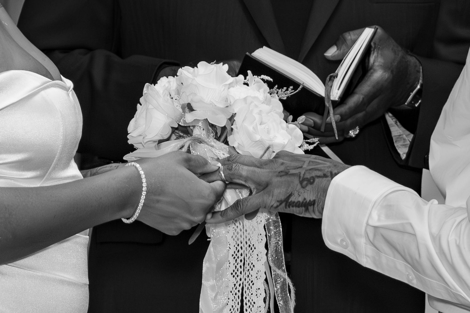 Black and white close-up of wedding ring exchange ceremony with bride holding a white floral bouquet.