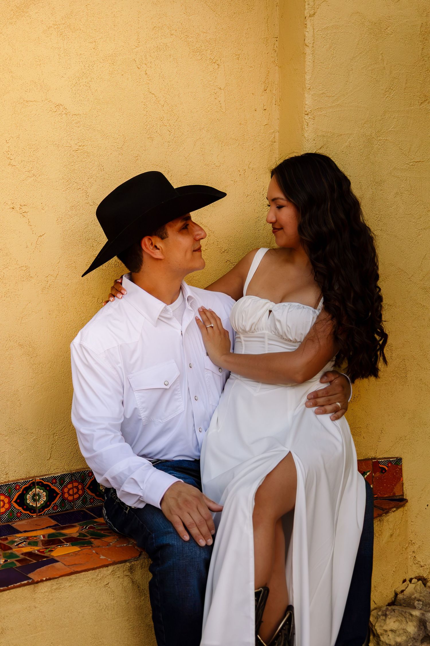 A couple in Western attire shares a romantic moment, man in black cowboy hat and woman in white dress, against a yellow wall.