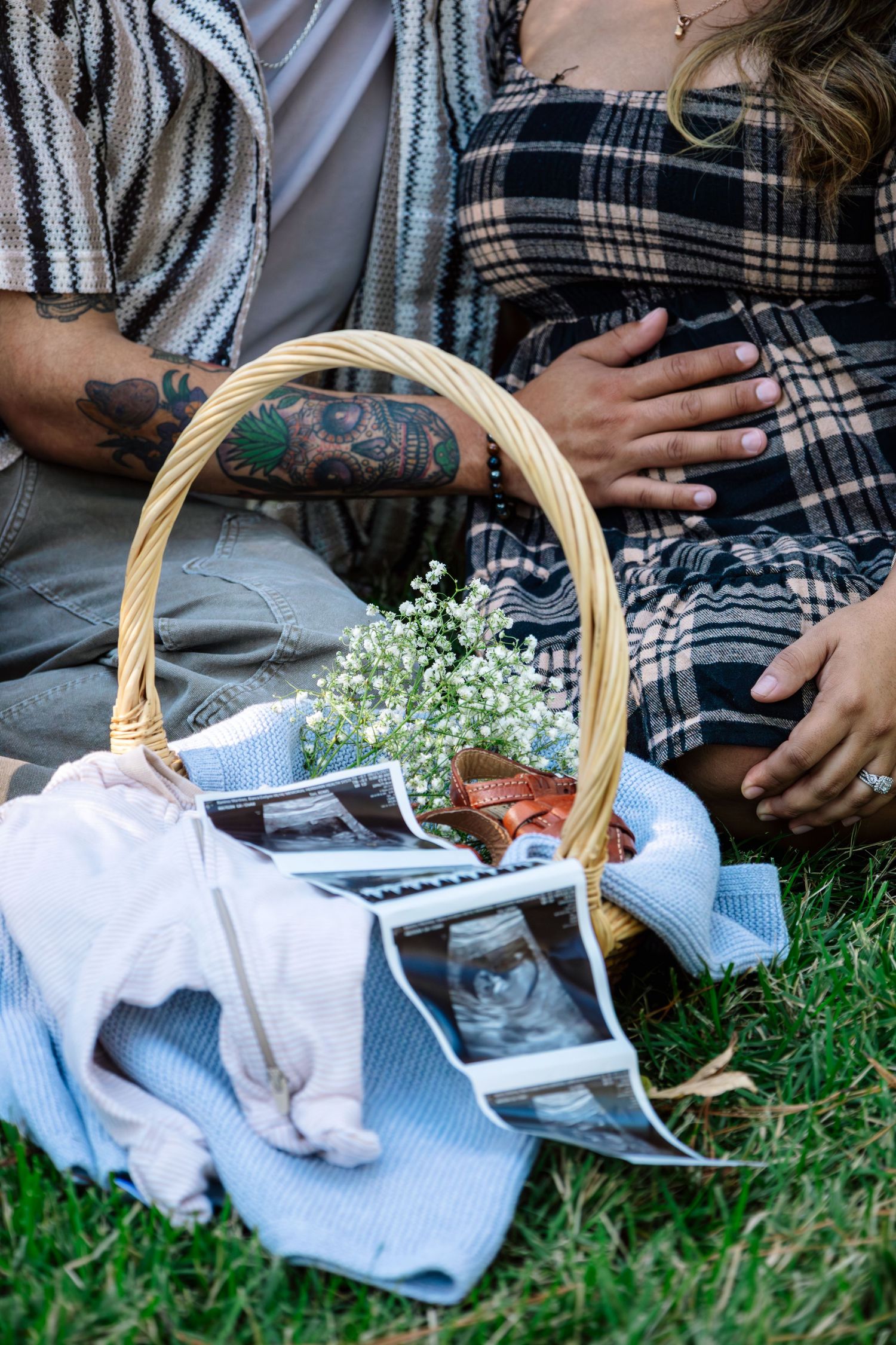 Couple sitting on grass with wicker basket containing ultrasound photos and baby clothes for pregnancy announcement.