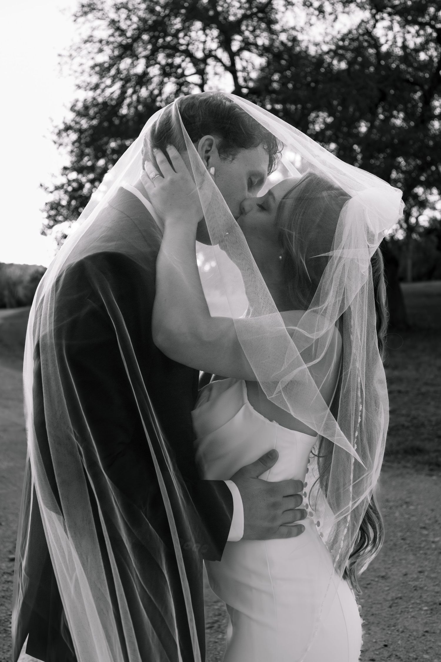 Black and white romantic photo of bride and groom kissing under a flowing wedding veil outdoors.