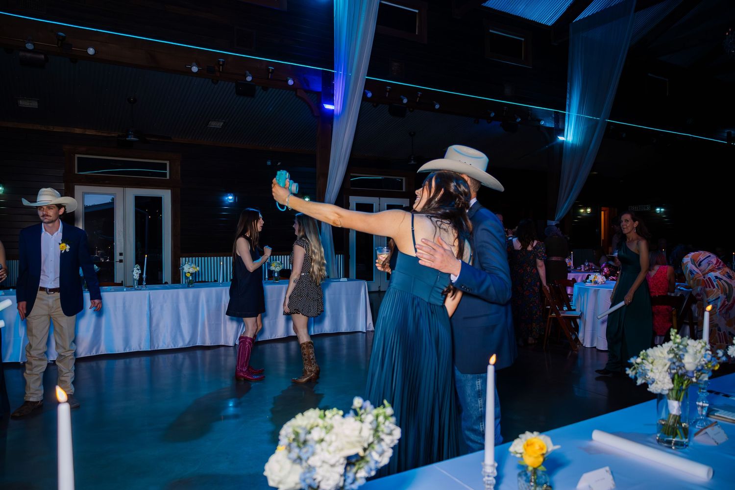 Guests in cowboy hats dancing at an elegant wedding reception with blue lighting and floral centerpieces.