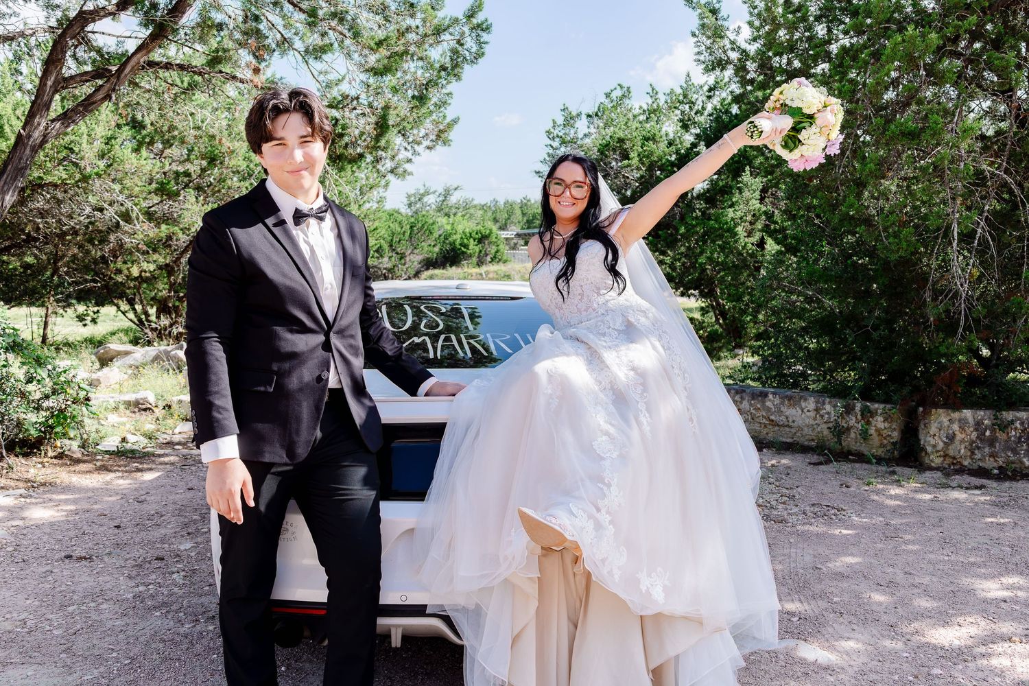 Bride and groom posing by a Just Married car outdoors, bride raising bouquet joyfully in wedding attire.