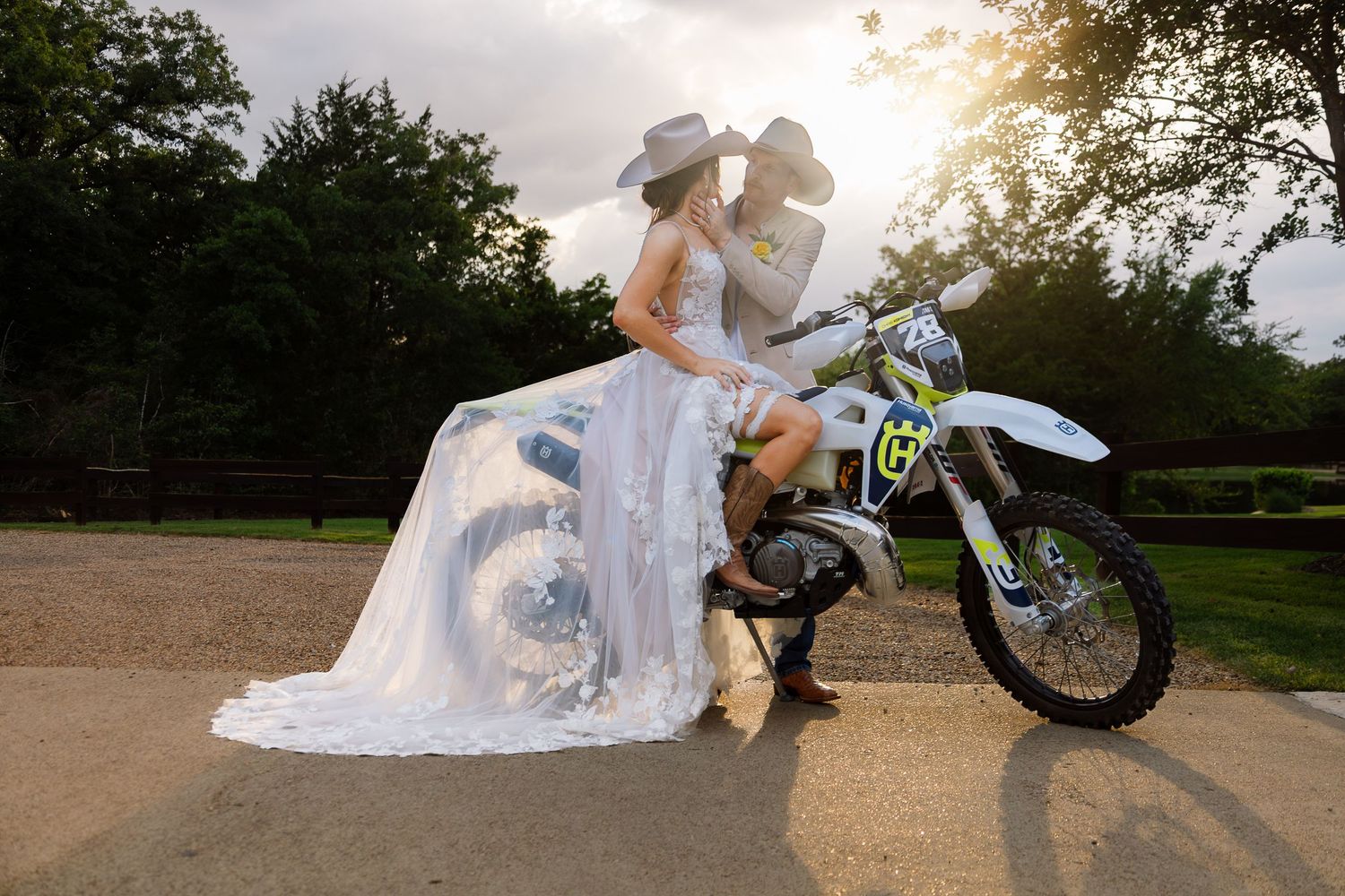 Bride and groom kissing on a dirt bike in wedding attire, with a flowing white gown and cowboy hat, at golden hour.