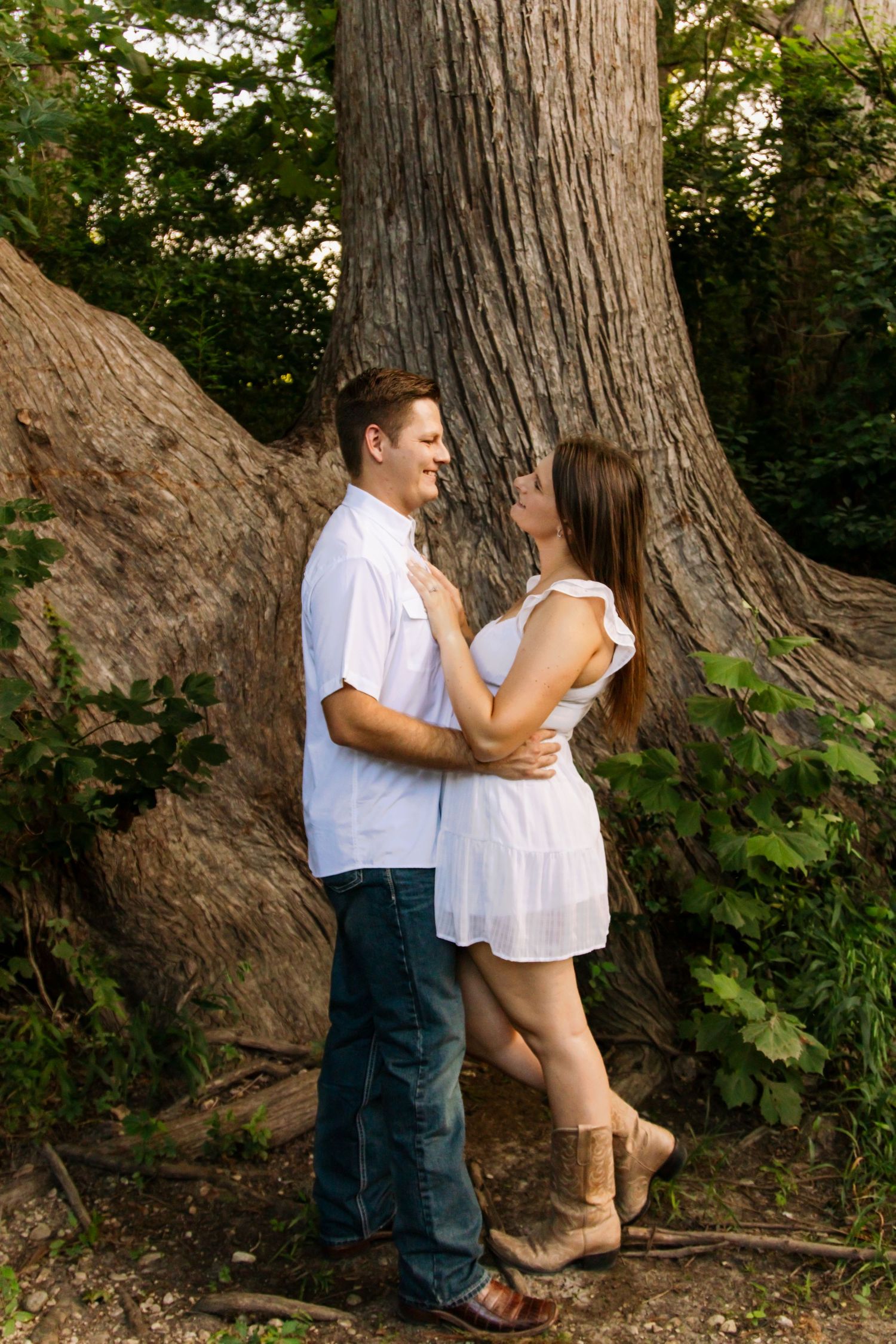 Couple sharing a romantic moment embracing in front of a large tree trunk in a wooded setting.