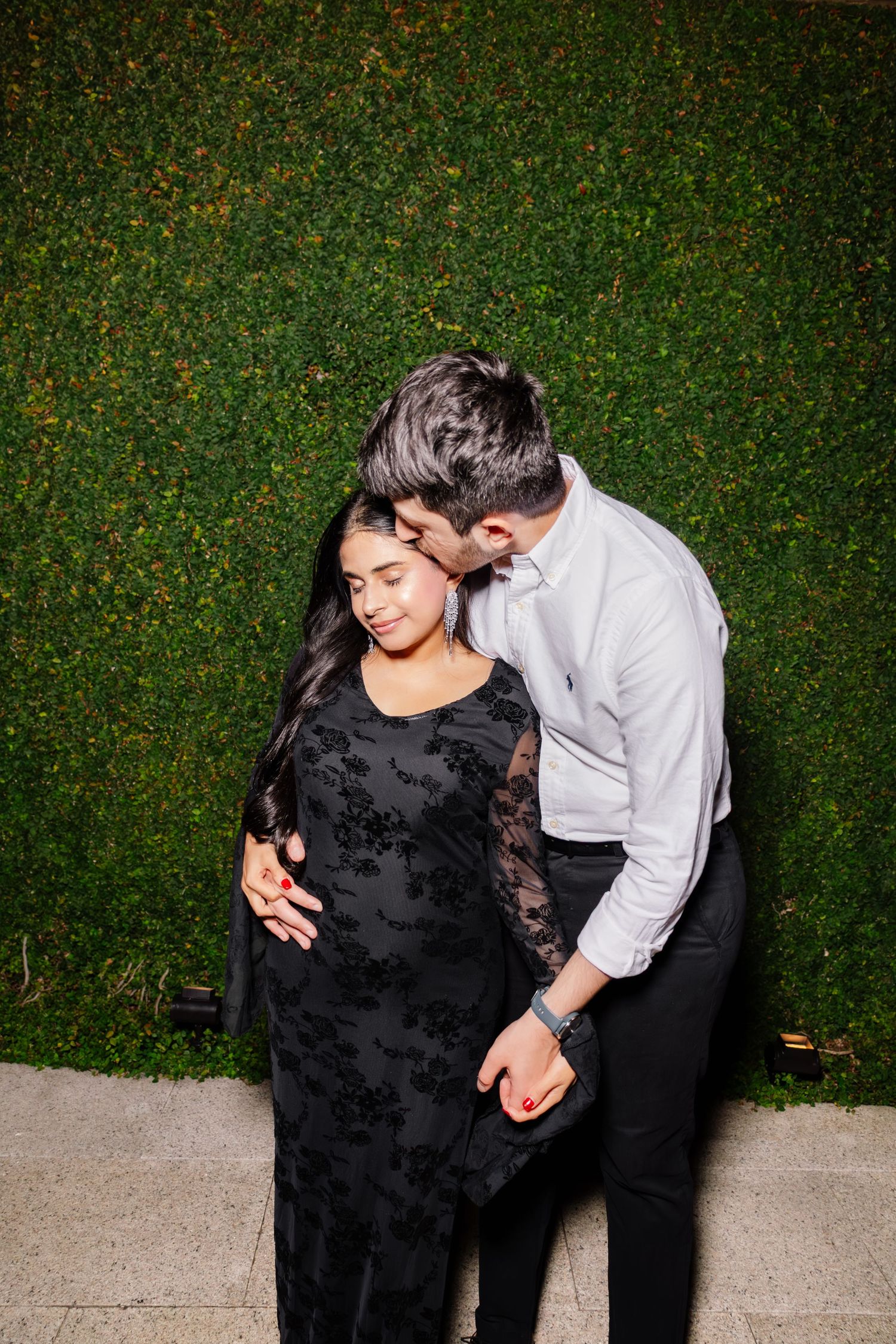 Couple shares an intimate embrace against a lush green hedge wall, woman wearing elegant black lace gown.