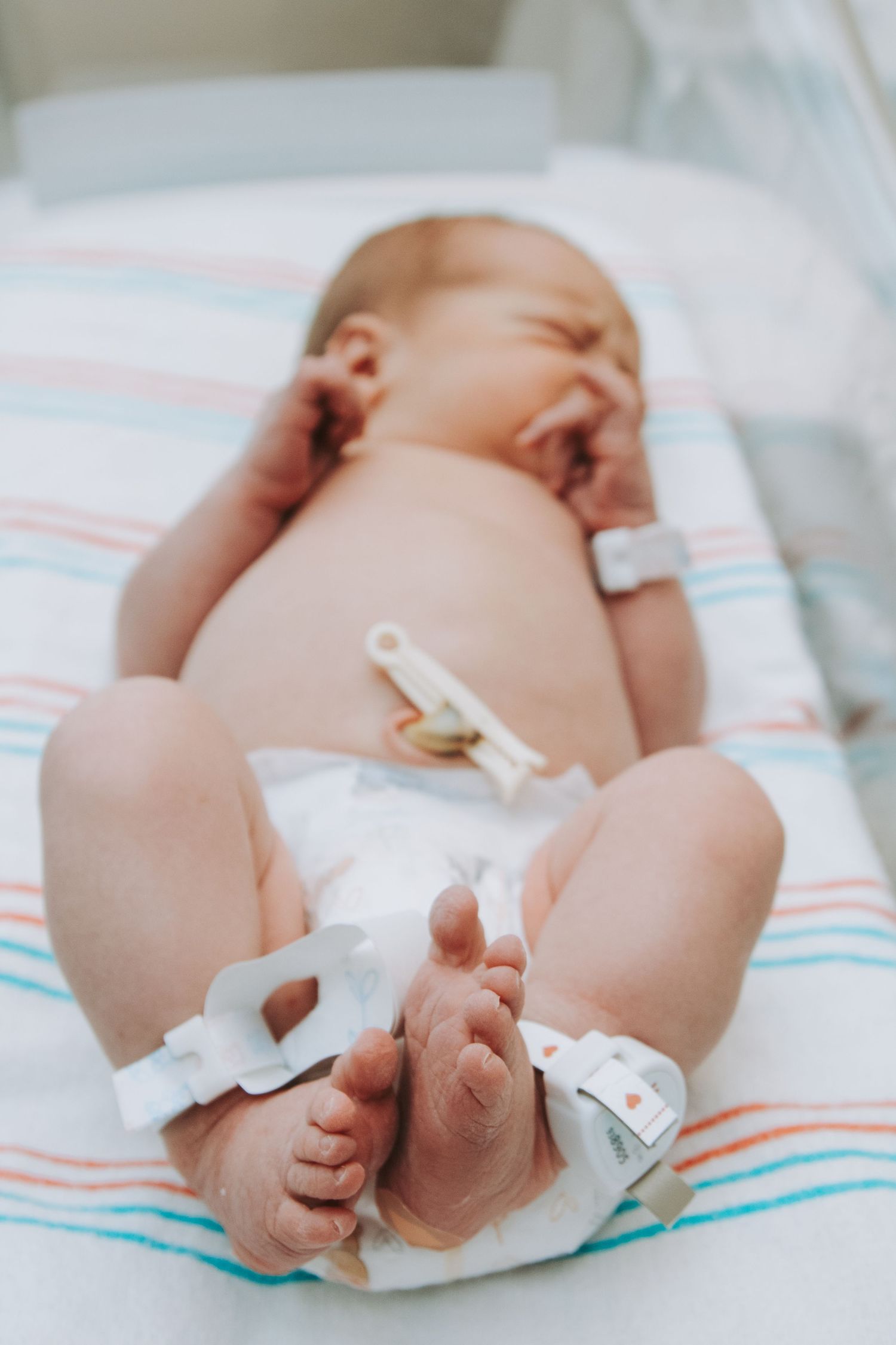 Newborn baby in a diaper lies on a striped hospital blanket with ID bands on wrists and ankles.
