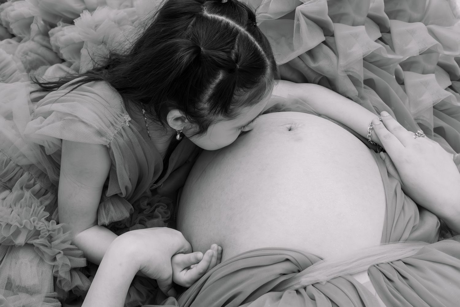 Black and white photo of a young girl kissing her mothers pregnant belly surrounded by tulle.