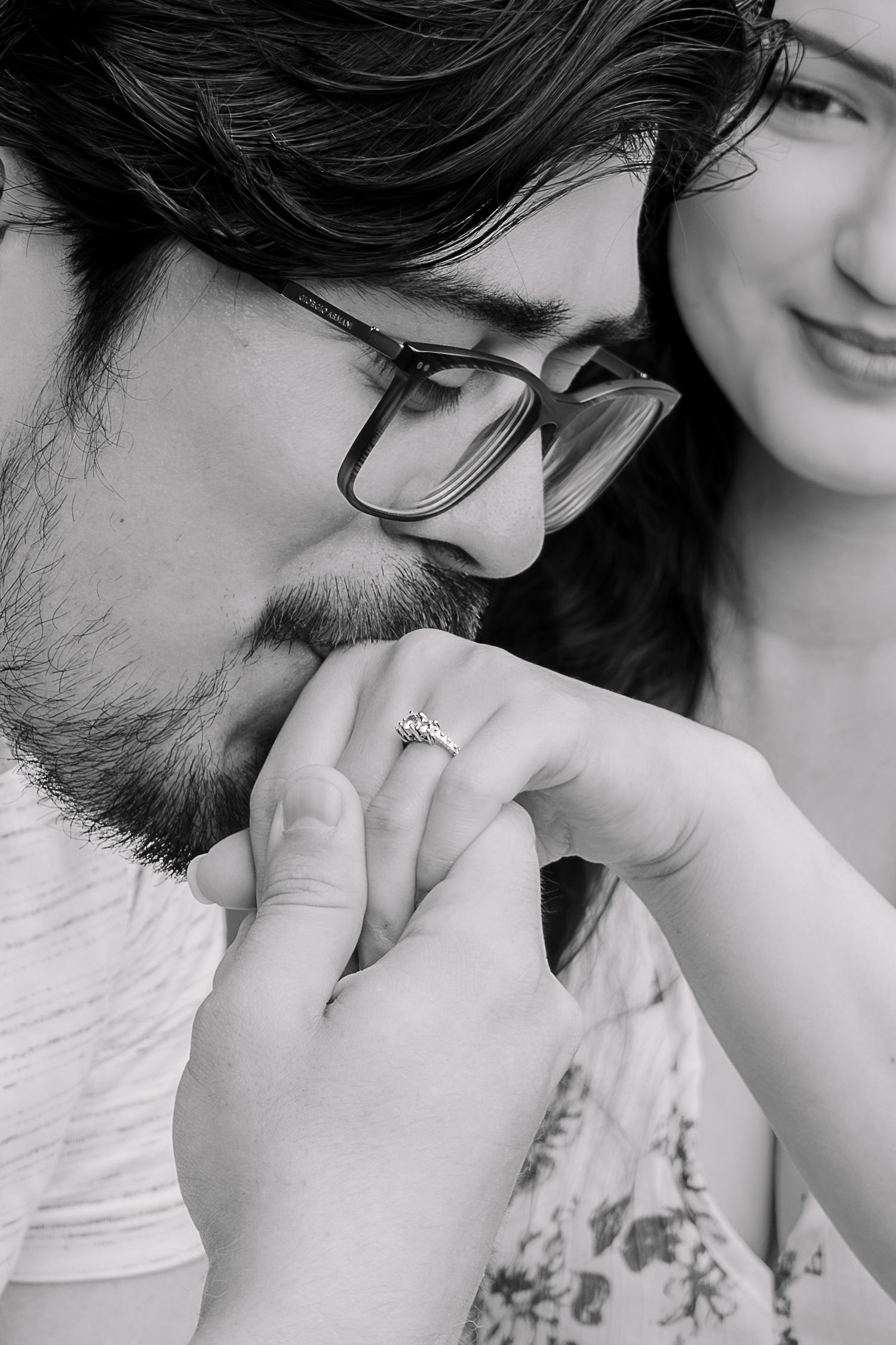 Black and white photo of a man kissing a woman's hand featuring an engagement ring while she smiles warmly.