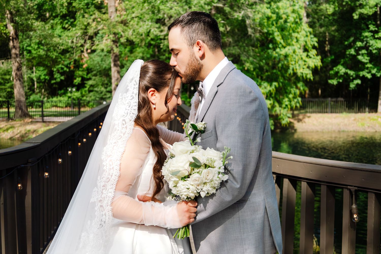 Bride and groom share a romantic moment on a wooden bridge surrounded by lush green trees at their outdoor wedding.