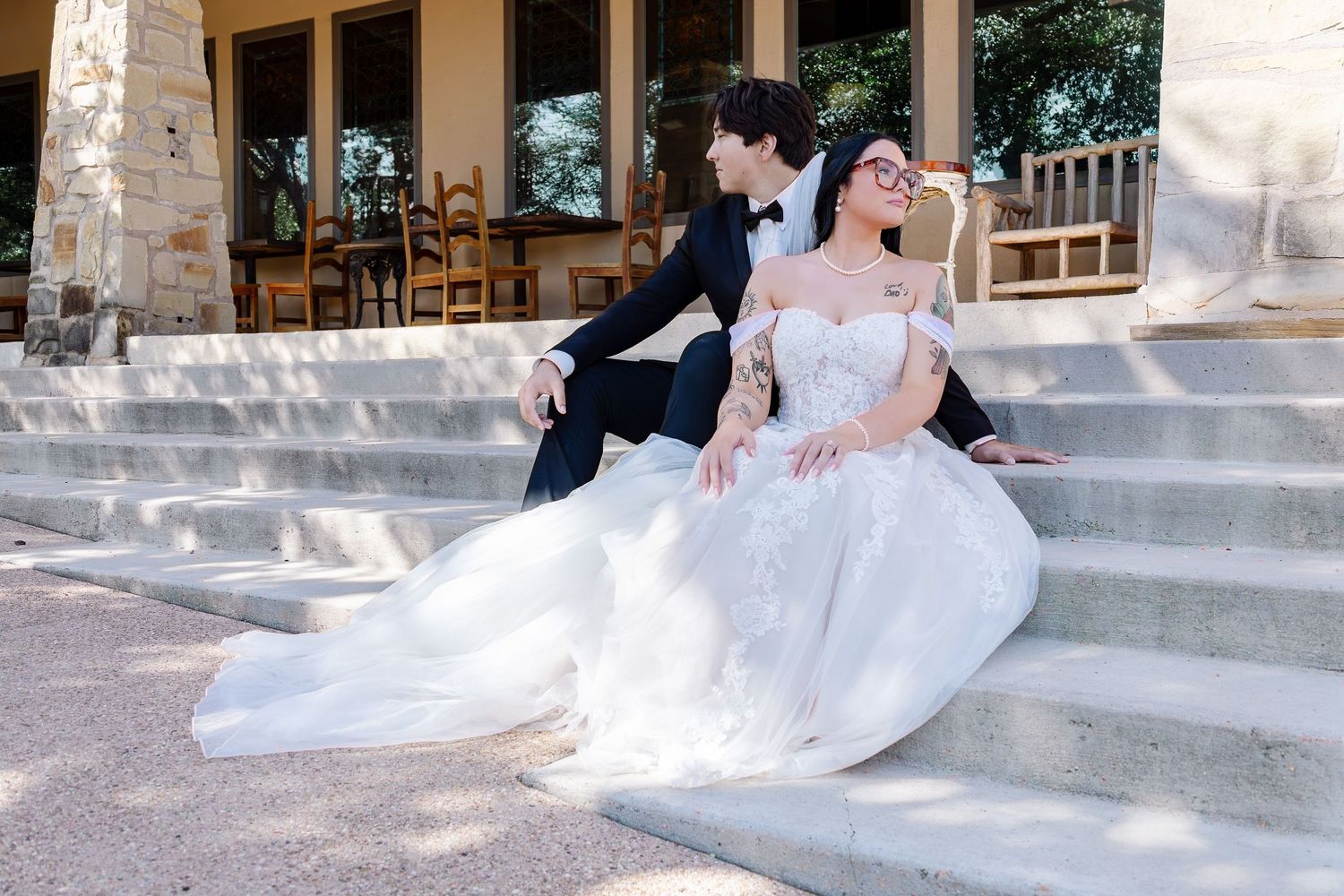 Bride in white gown and groom in navy suit sit together on stone steps at an outdoor wedding venue.