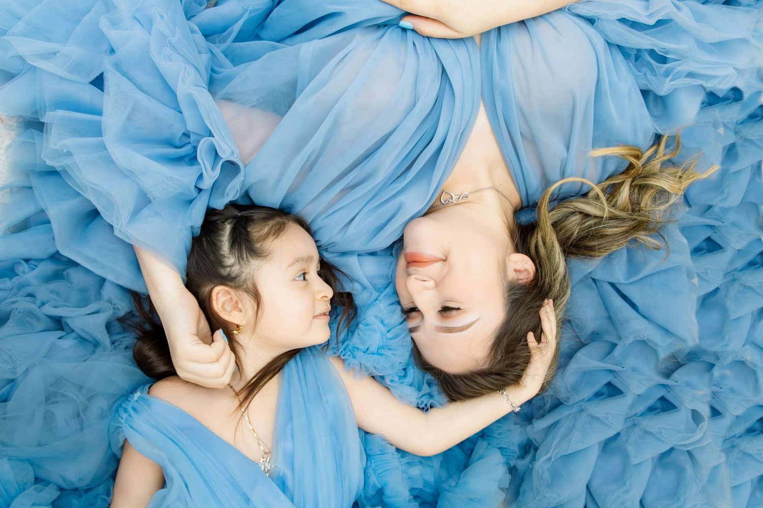 Mother and daughter in matching blue tulle gowns lie on the floor, smiling at each other.