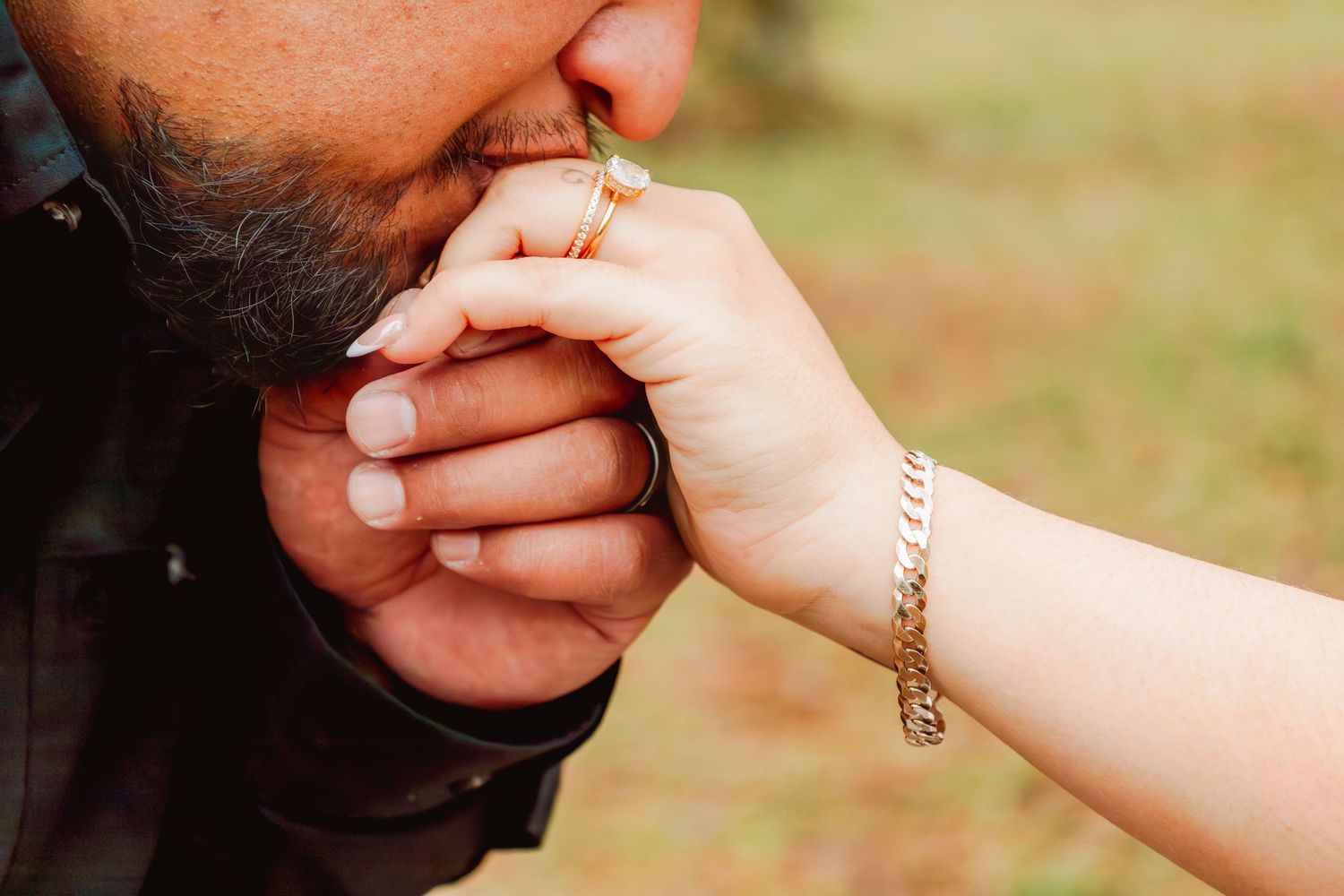 Groom tenderly kissing bride's hand adorned with a gold bracelet and ring outdoors.